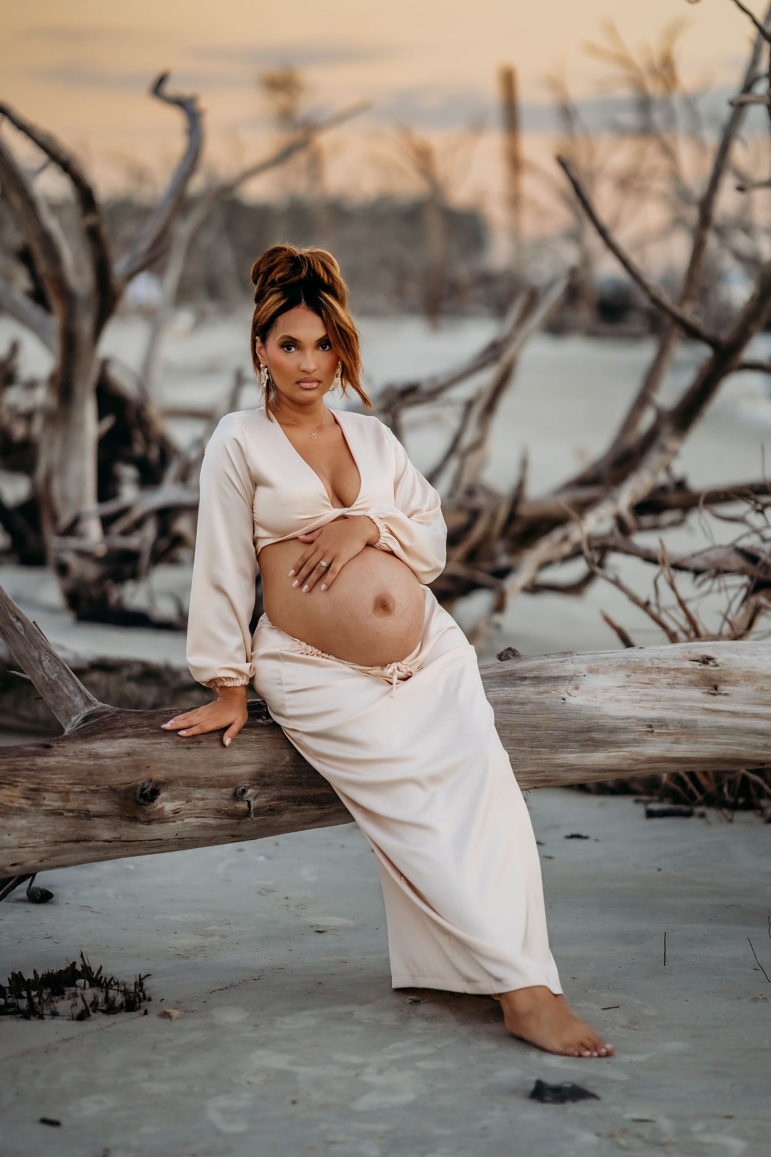 Pregnant woman in cream two-piece outfit poses against driftwood at sunset beach with serene expression.