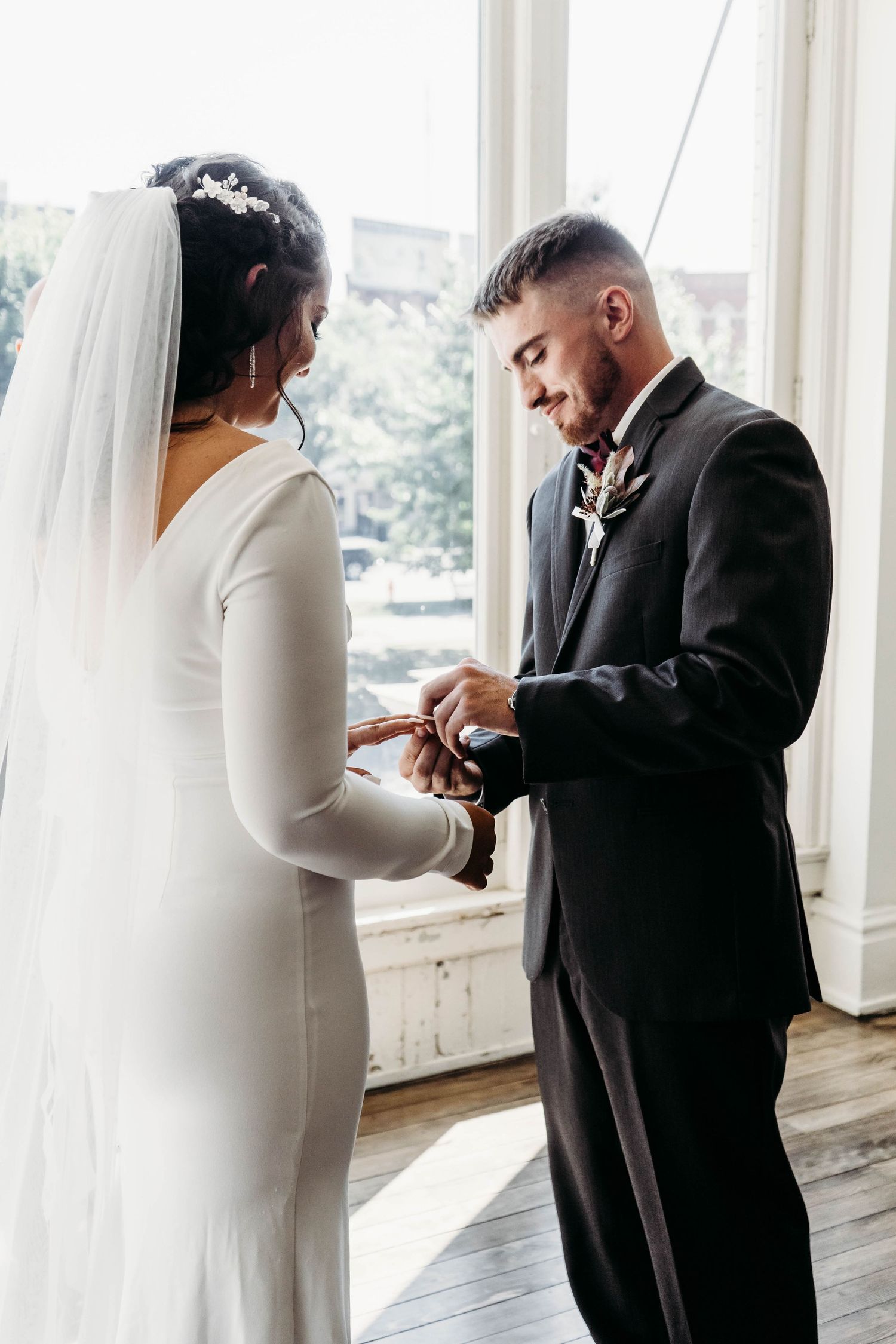 Groom in black suit placing ring on brides finger during indoor wedding ceremony.