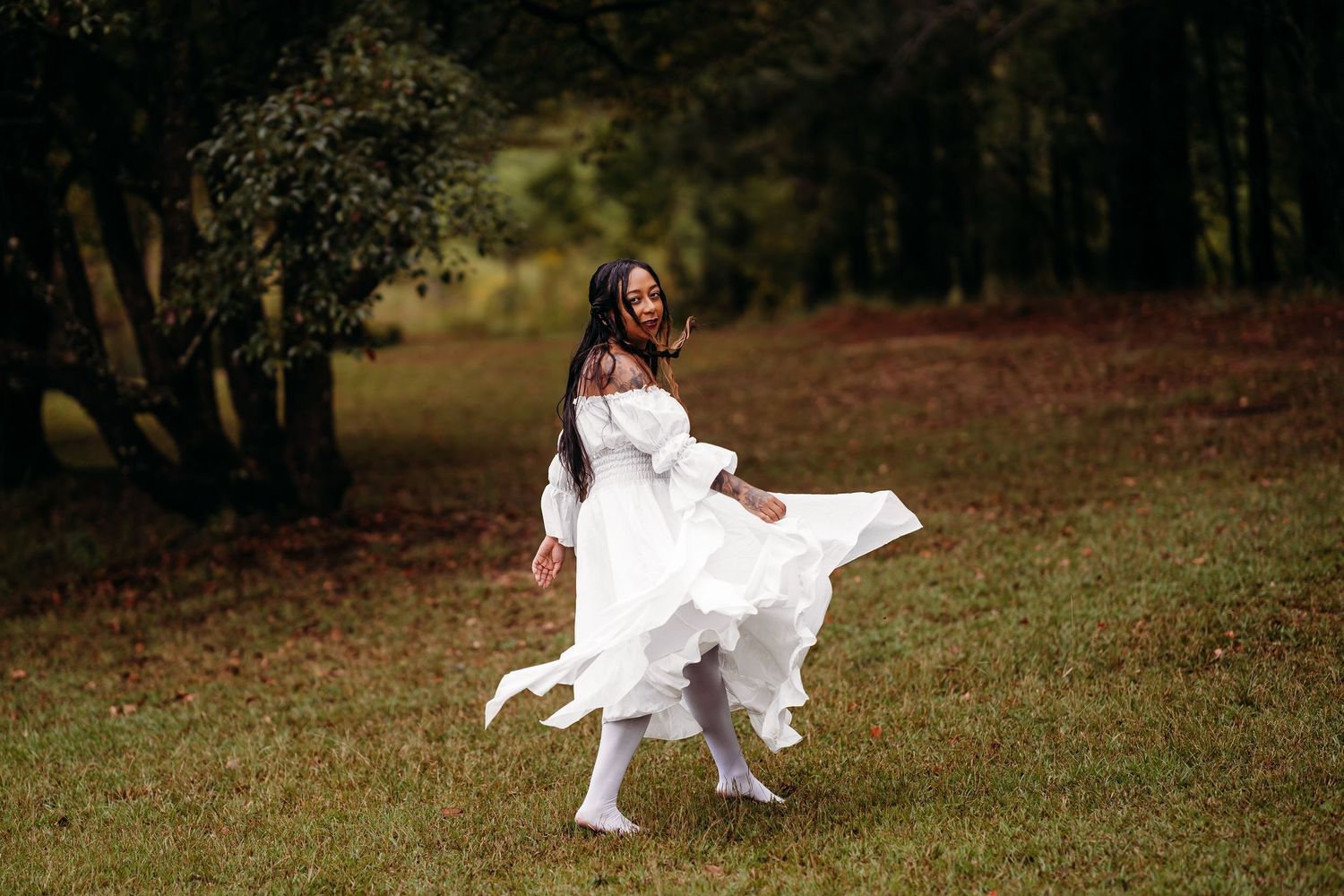 Woman in white off-shoulder dress twirling gracefully on green grass with forest backdrop.