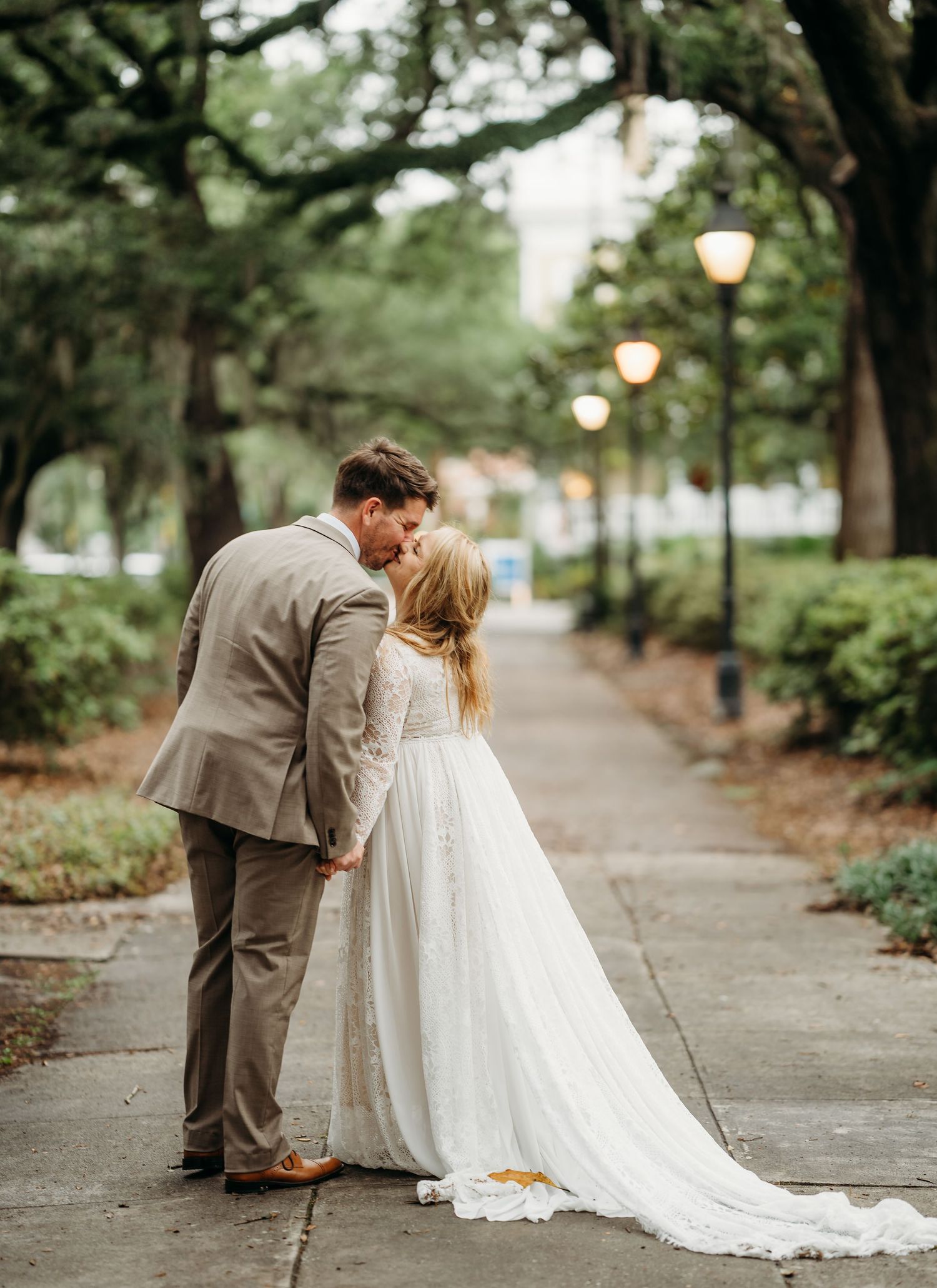Bride and groom share a tender kiss on a picturesque tree-lined path illuminated by vintage glowing street lanterns.