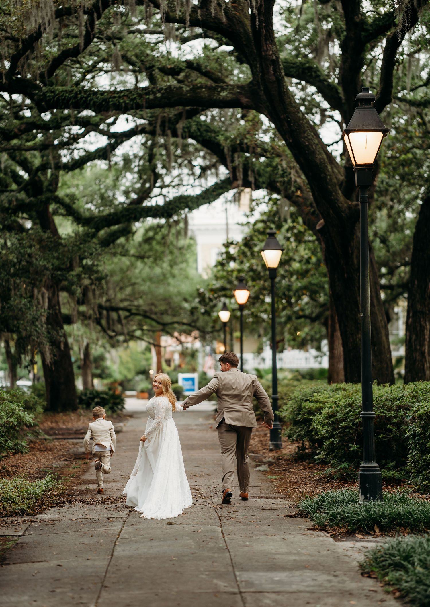 Bride and groom hold hands walking a tree-canopied path with a child running ahead beneath glowing vintage street lamps.