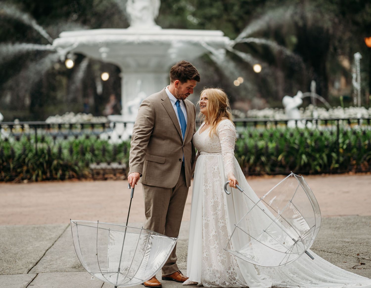 Bride and groom smile at each other holding clear umbrellas with white park fountain in background.