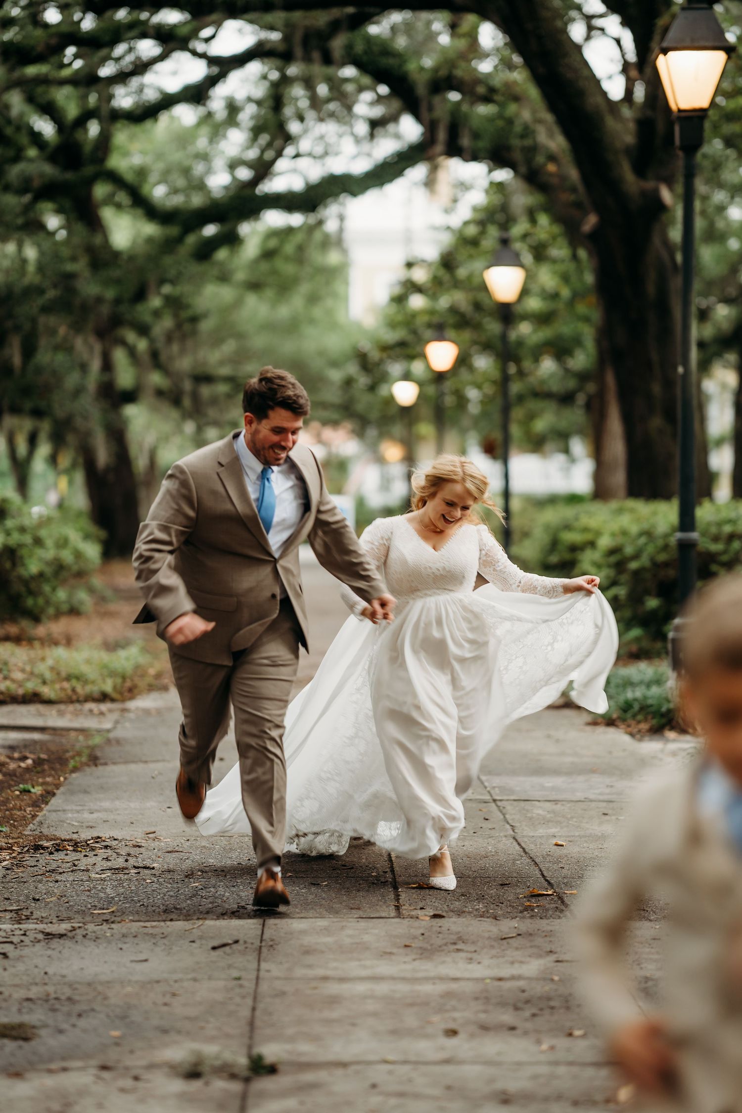 Laughing bride and groom run playfully along lamp-lit park pathway in wedding attire.