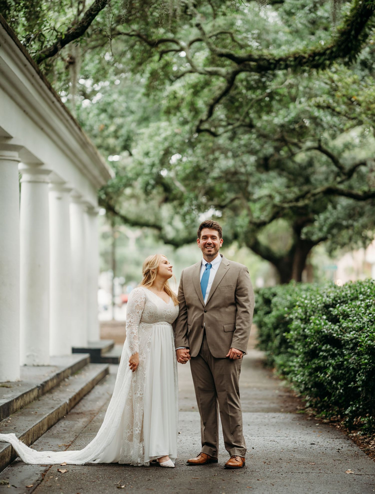 Bride and groom holding hands on a tree-lined pathway with white columns at an outdoor wedding venue.
