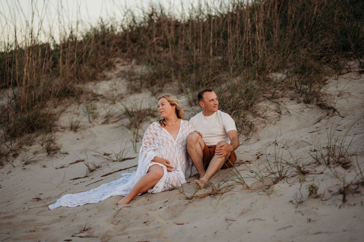 Couple sitting together on sandy beach dunes at sunset, woman in white dress, man in white shirt, surrounded by sea grass.