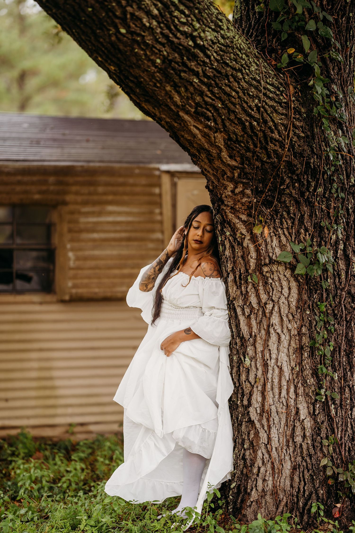 Woman in white off-shoulder dress leaning against large tree near rustic wooden structure.