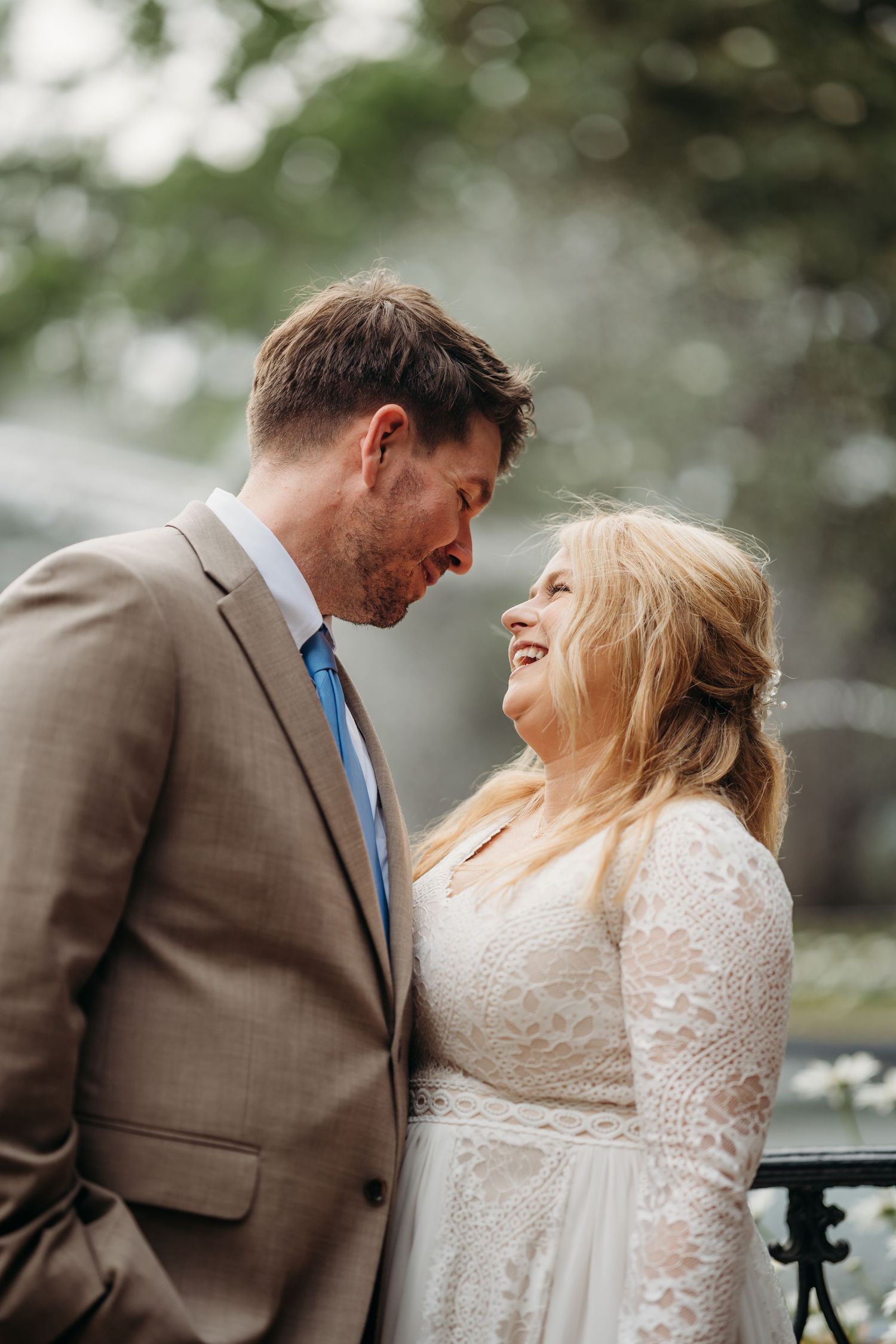 Bride laughs joyfully as groom gazes at her lovingly, both standing close together in an outdoor garden setting.