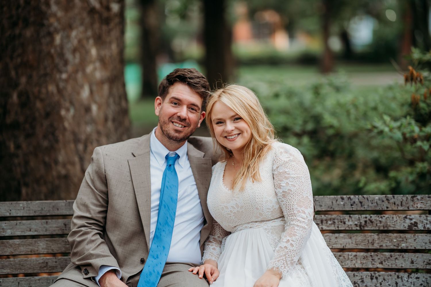 Happy newlyweds smiling at the camera while seated on a park bench on their wedding day.