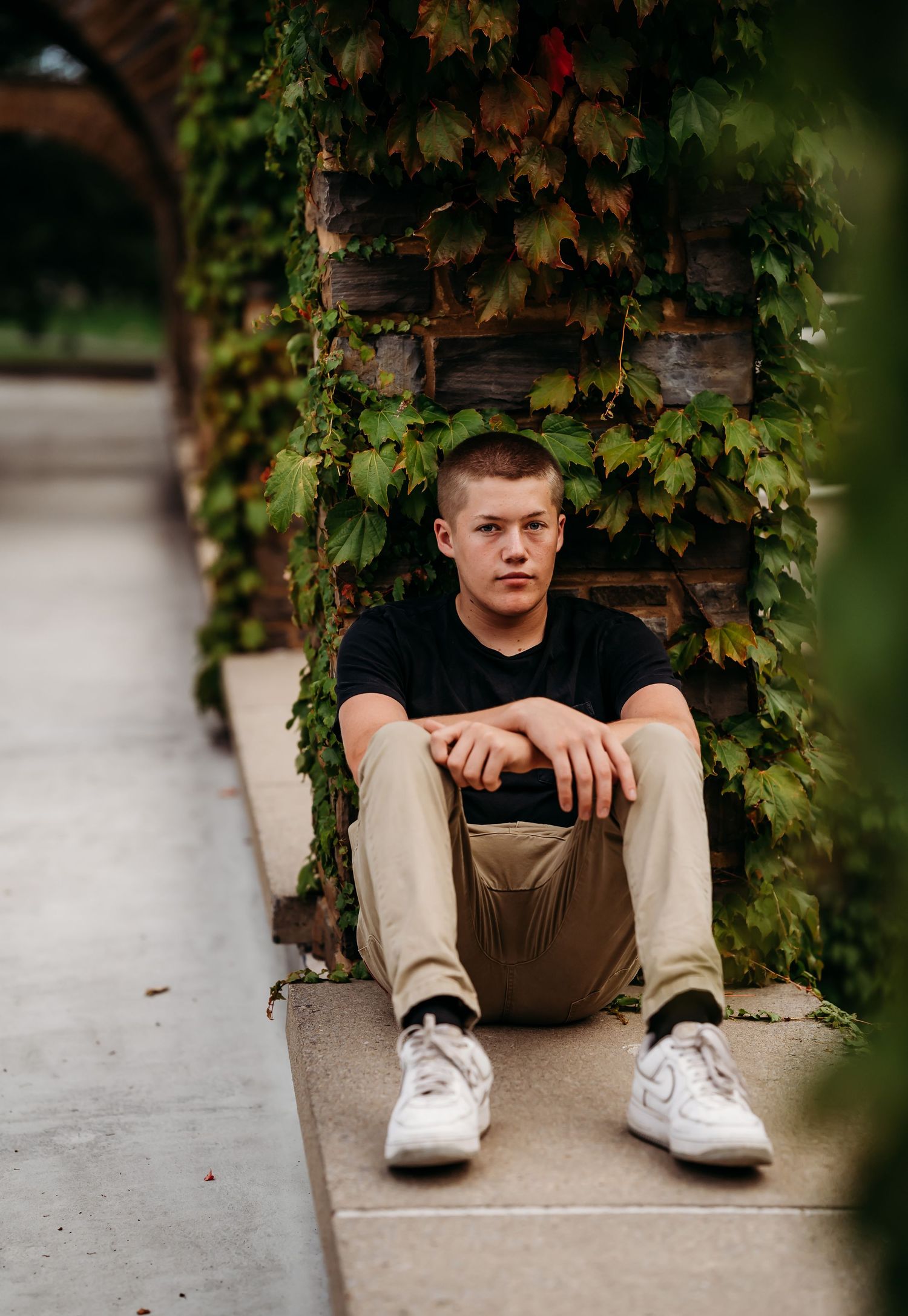 Young man in black shirt and khakis sitting on steps beside ivy-covered brick archway.