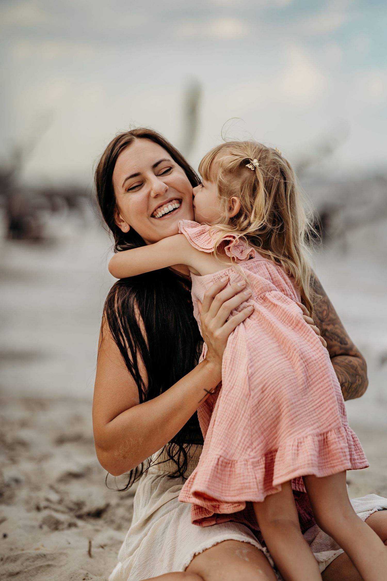 A laughing mother holds her young daughter in a pink dress on a sandy beach, sharing a sweet kiss.