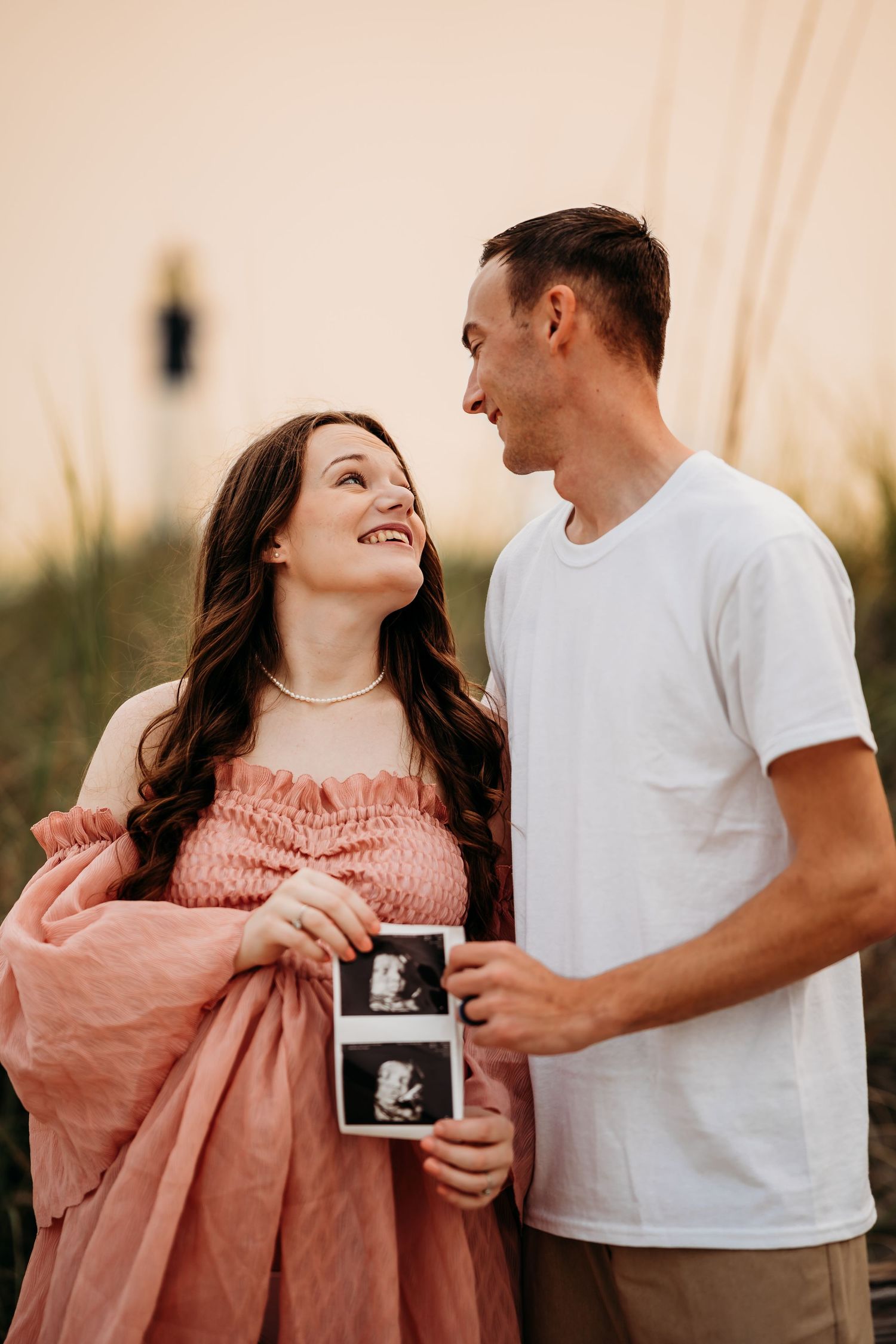 Happy expecting couple gazing at each other while holding ultrasound photos on beach at dusk.