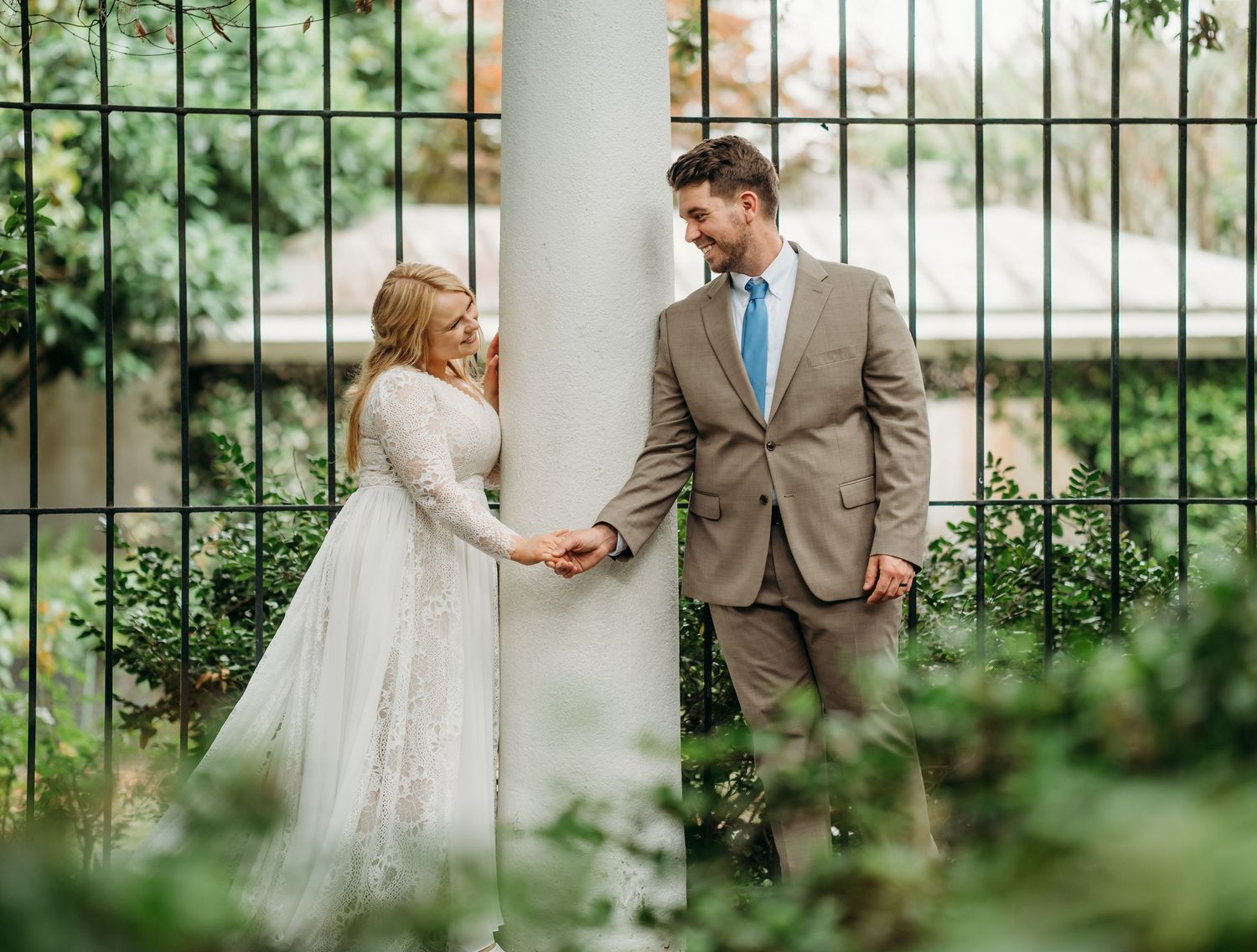 Bride and groom holding hands and smiling at each other beside a white column with green foliage backdrop.