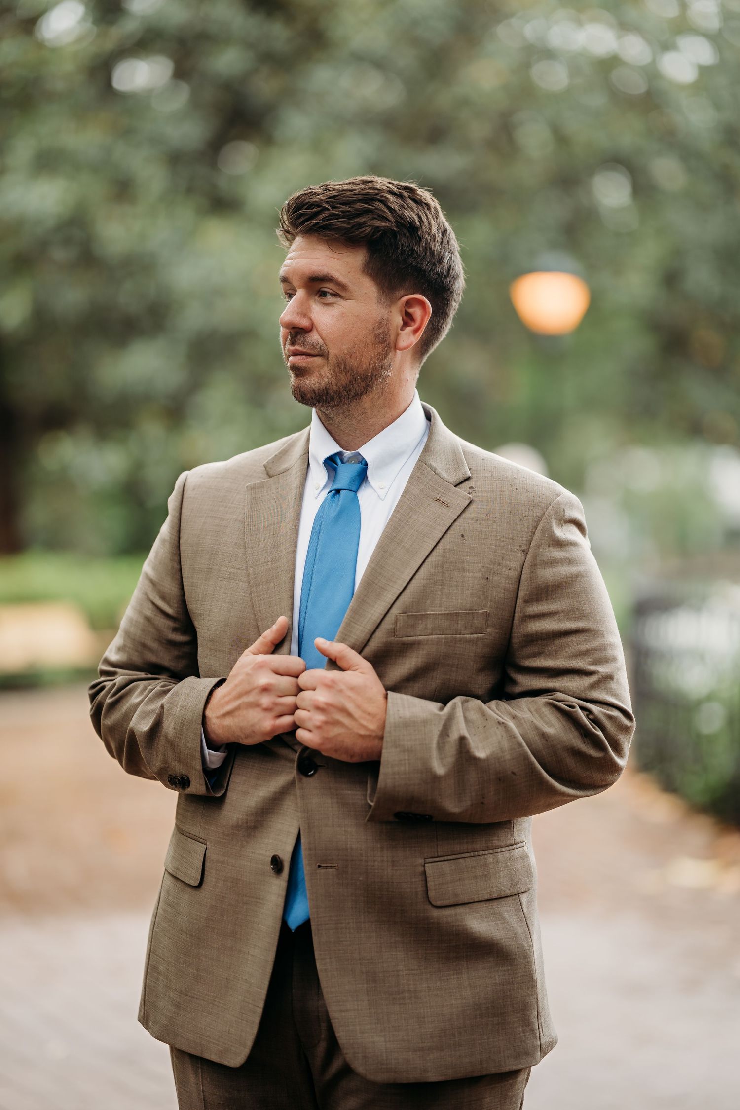 Groom in tan suit and blue tie adjusting jacket outdoors in park setting.
