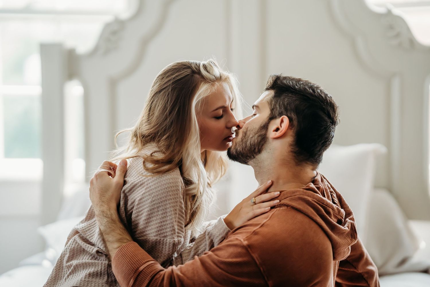 Couple sharing a romantic kiss while sitting on a bed in a bright bedroom.
