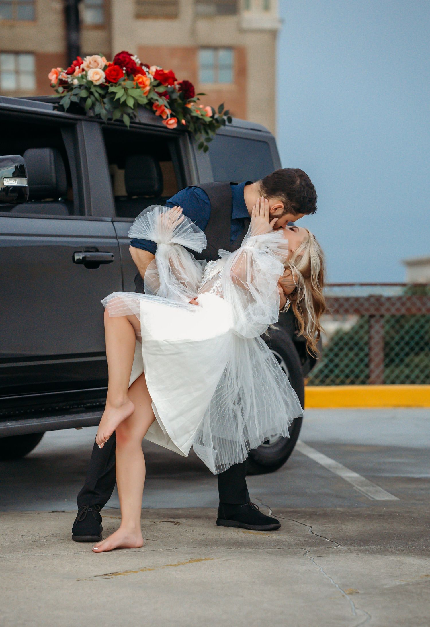 Groom dips bride in white dress for a romantic kiss beside a black SUV decorated with red floral arrangement.