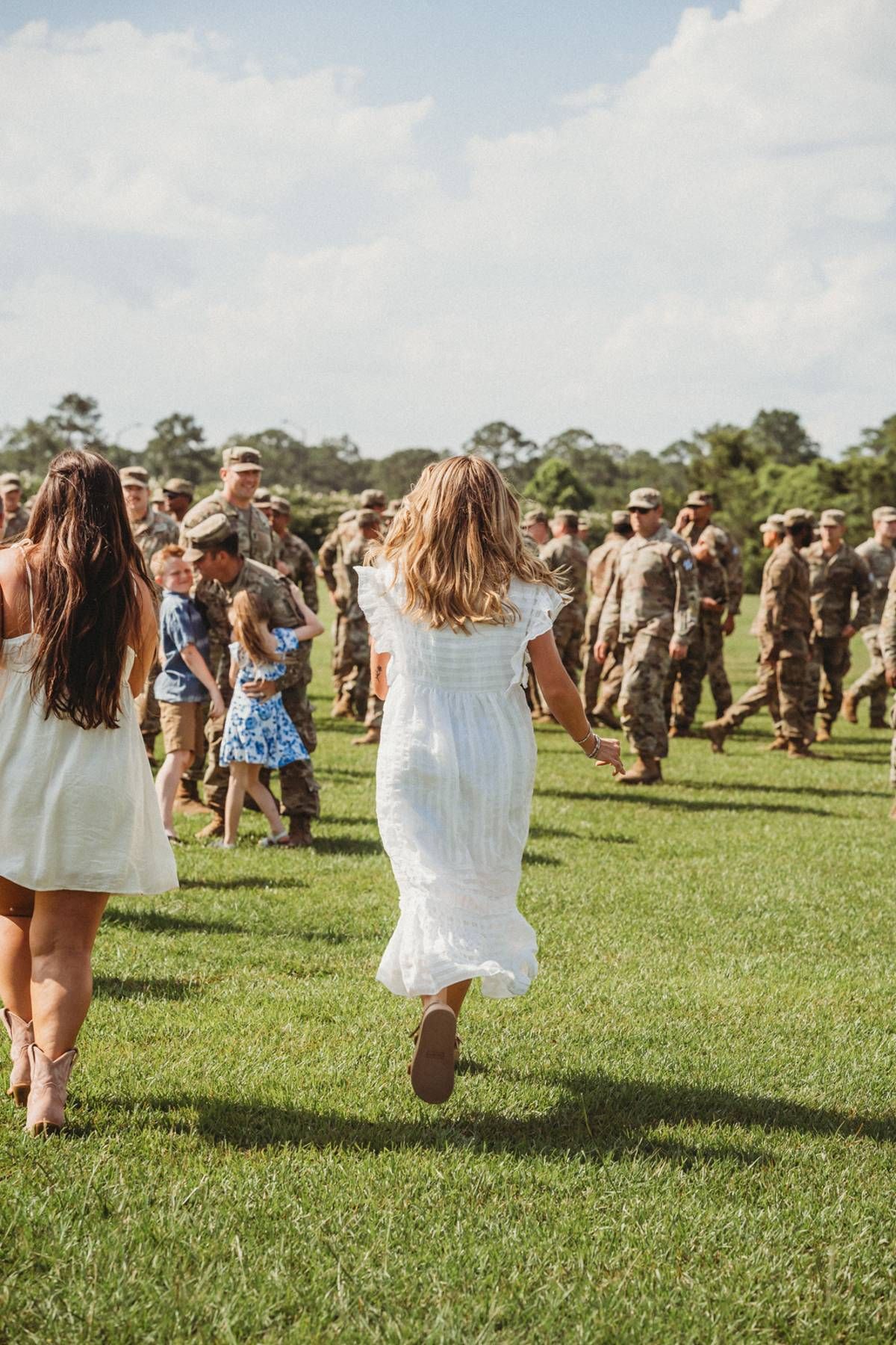 Women in white dresses run across green field toward military soldiers during homecoming reunion ceremony outdoors.