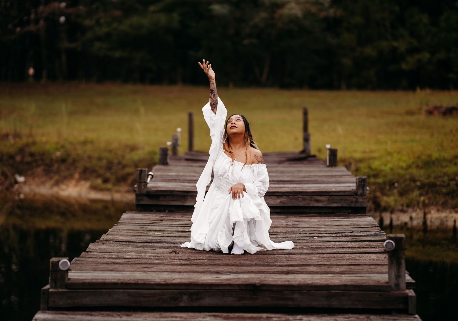 Woman in white off-shoulder gown poses gracefully on a wooden dock with arm raised skyward.