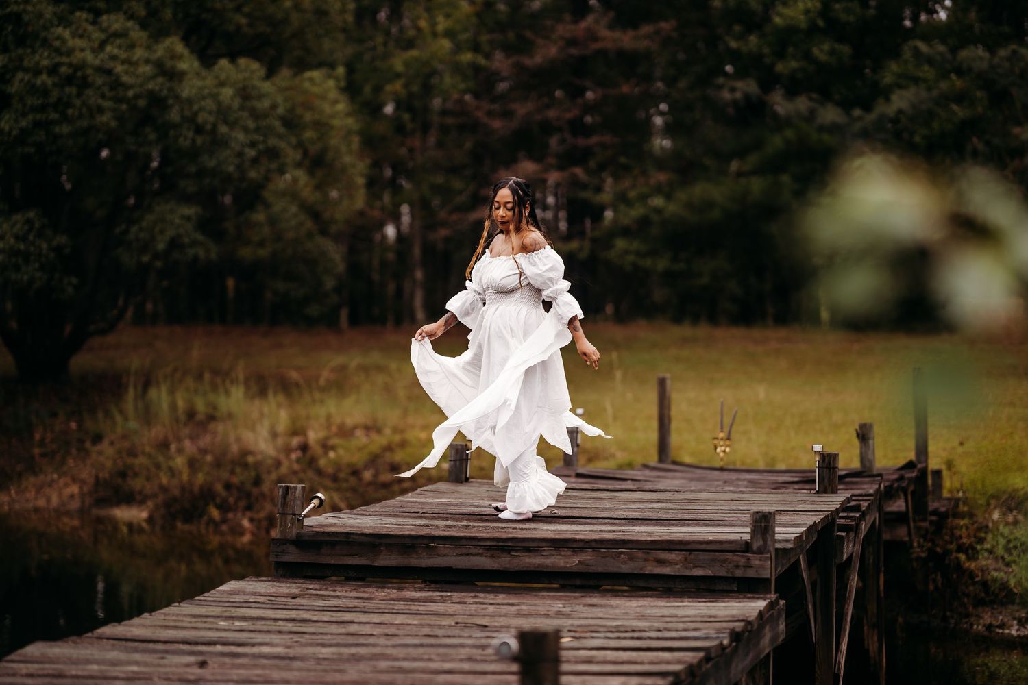 Woman in white off-shoulder dress dancing playfully on wooden dock with green forest background.