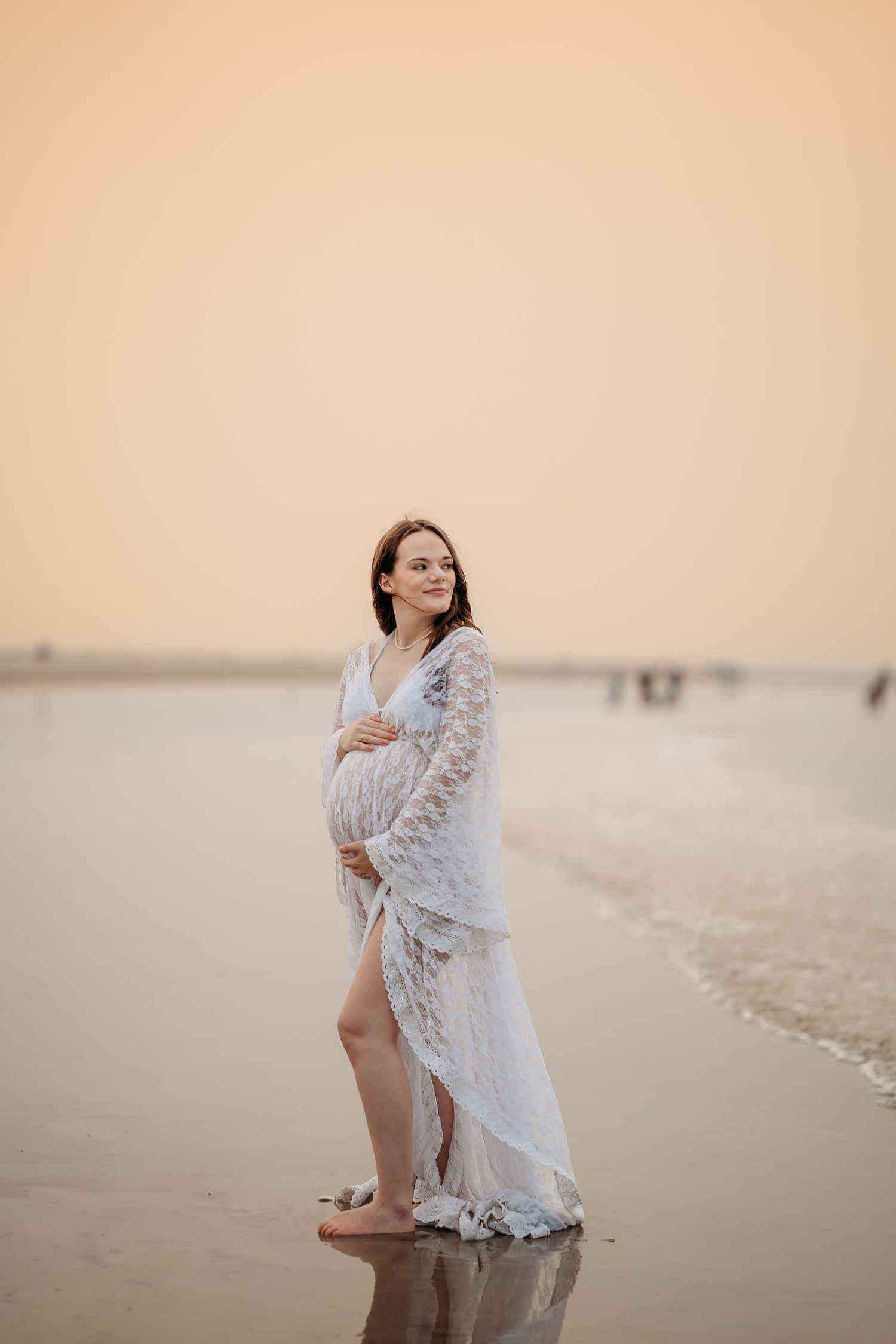 Pregnant woman in white lace dress standing at water's edge during golden sunset on beach.
