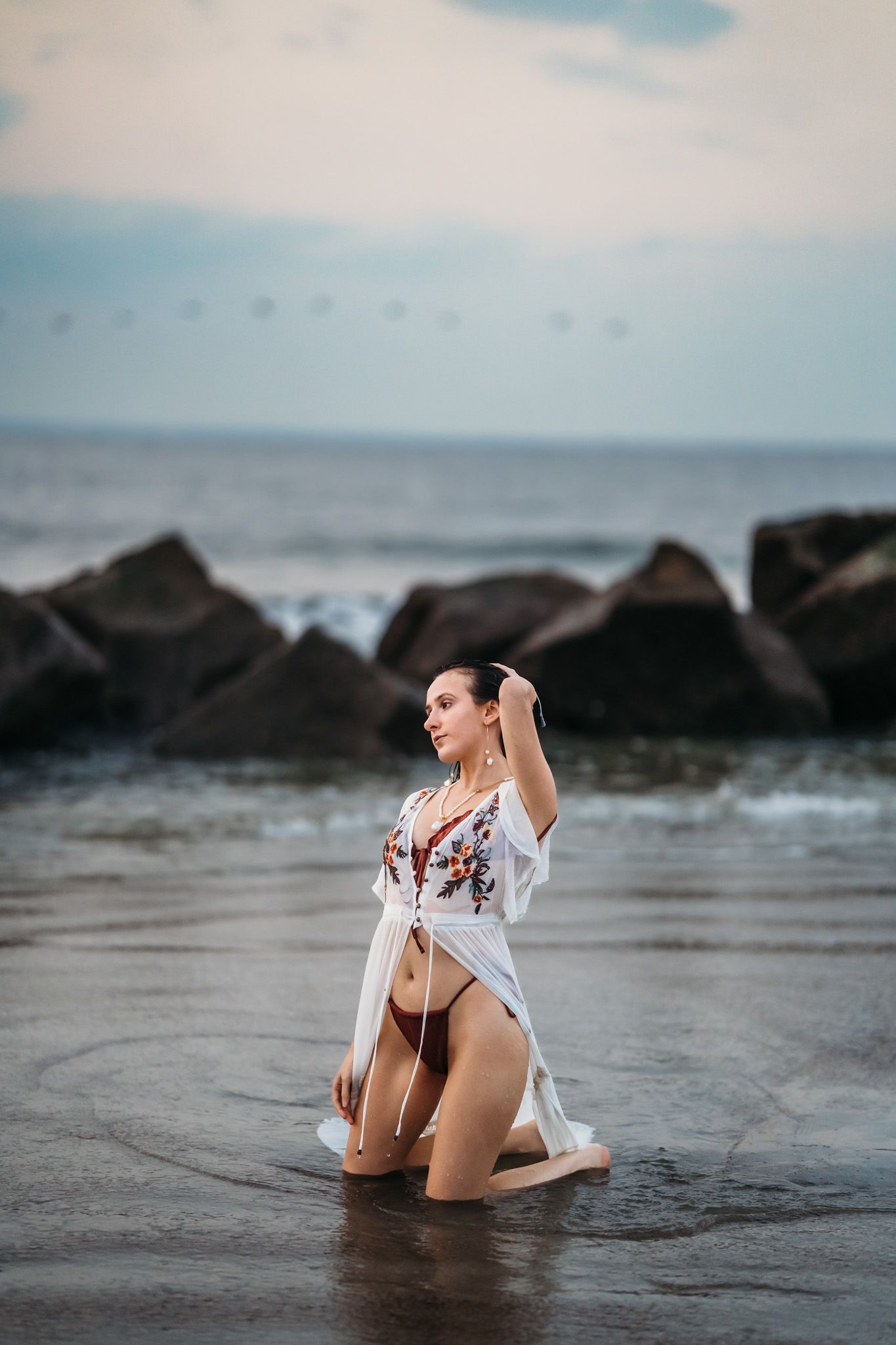 Woman in white floral kimono and bikini kneeling in ocean water at sunset with rocky shoreline backdrop.