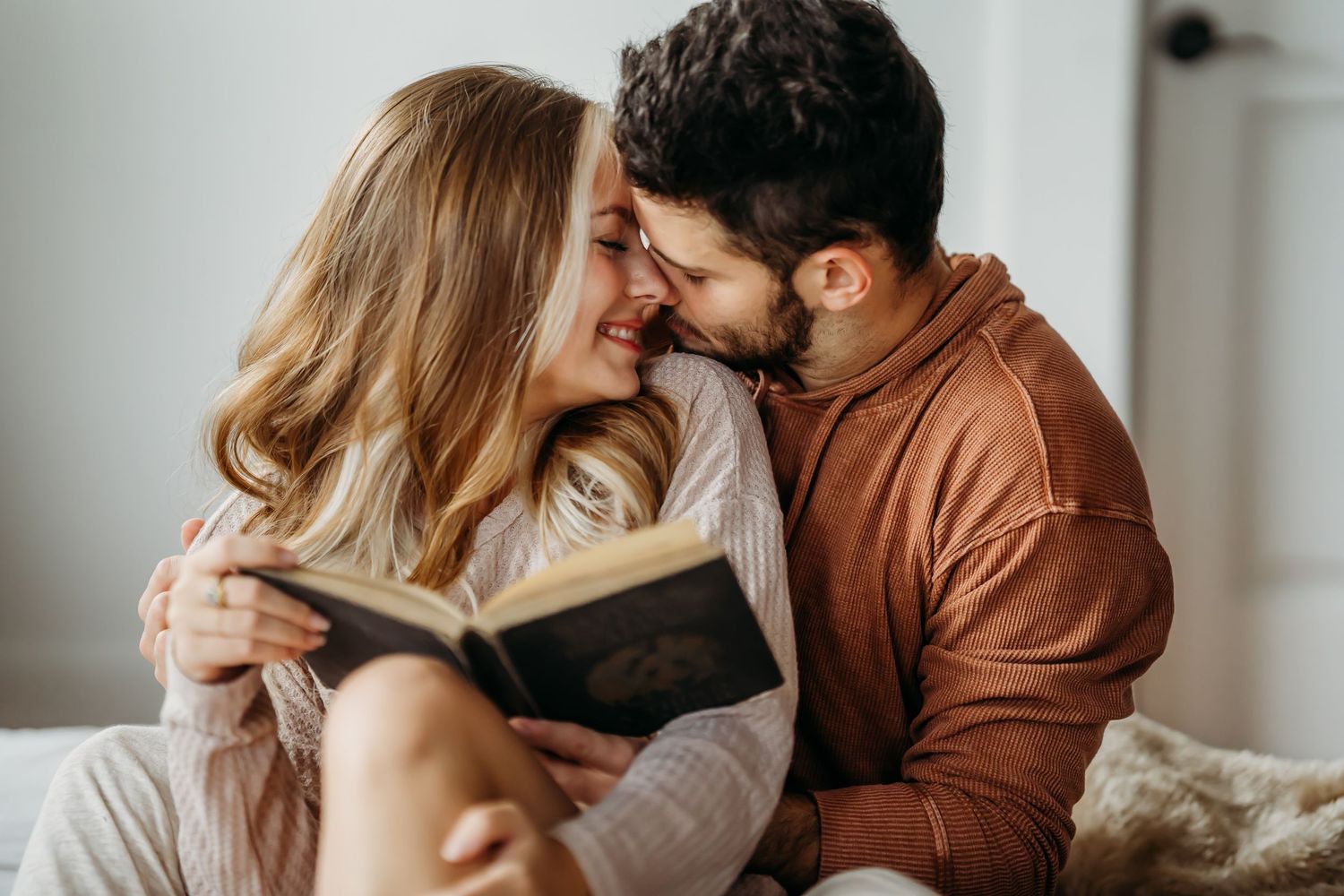 A smiling couple shares a tender moment while reading a book together on a cozy bed.