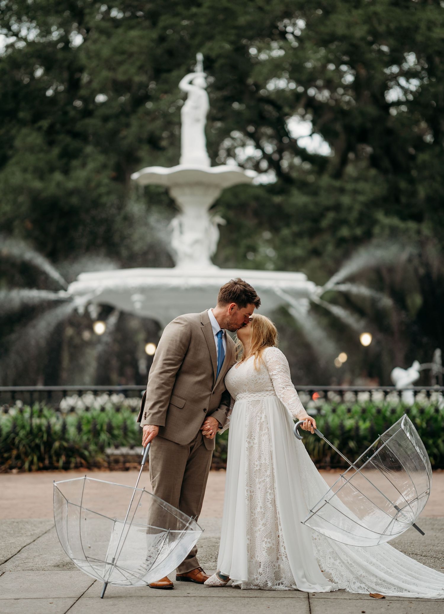 Bride and groom share romantic kiss holding clear umbrellas before grand white fountain in park setting.