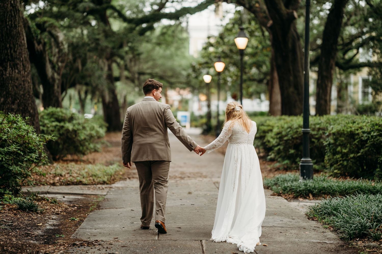 Bride in flowing white gown and groom in tan suit walk hand-in-hand down a lush, lamp-lit tree-canopied pathway.