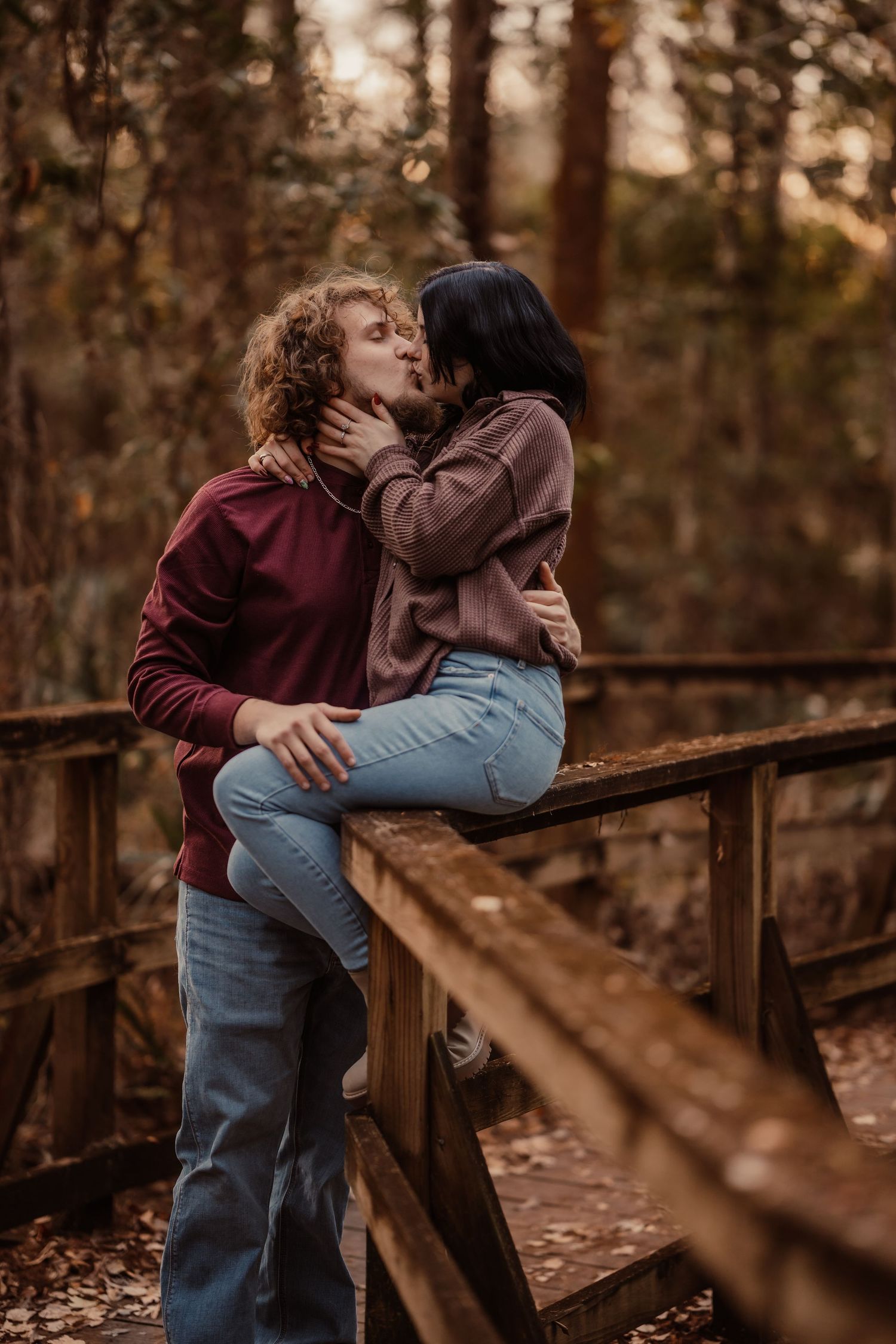 Couple sharing a romantic kiss on a wooden bridge railing surrounded by autumn forest foliage.