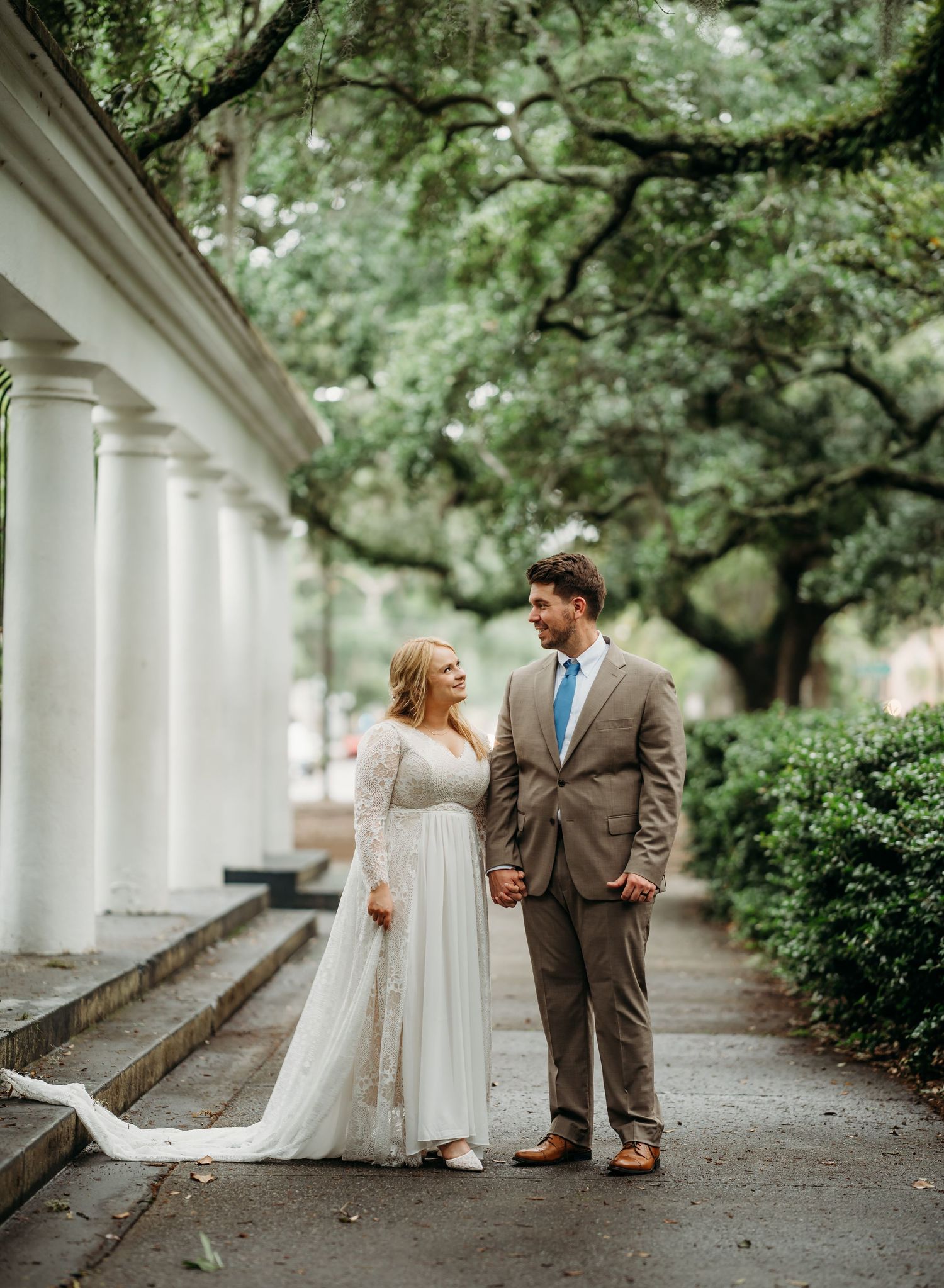 Bride in lace gown and groom in tan suit hold hands under oak trees by white columns.