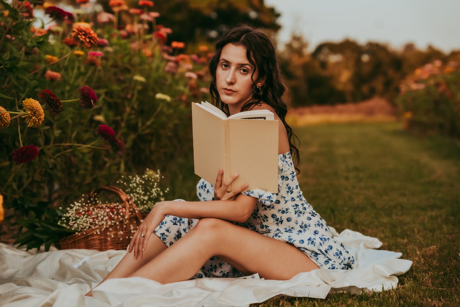 Young woman in floral dress reading a book outdoors on a blanket surrounded by colorful garden flowers at sunset.