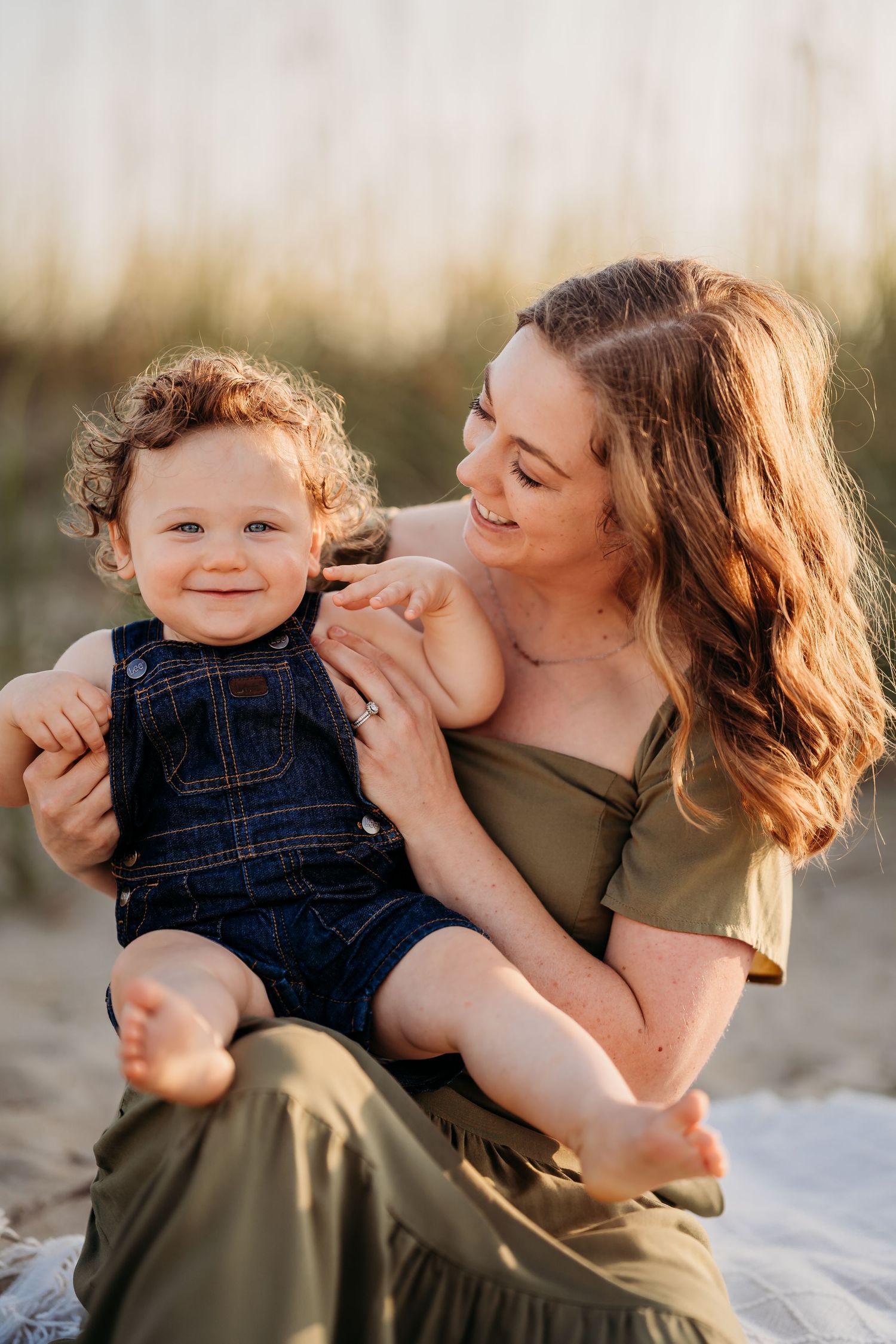 A smiling mother holds her happy toddler boy on a sandy beach at golden hour, both barefoot and casually dressed.
