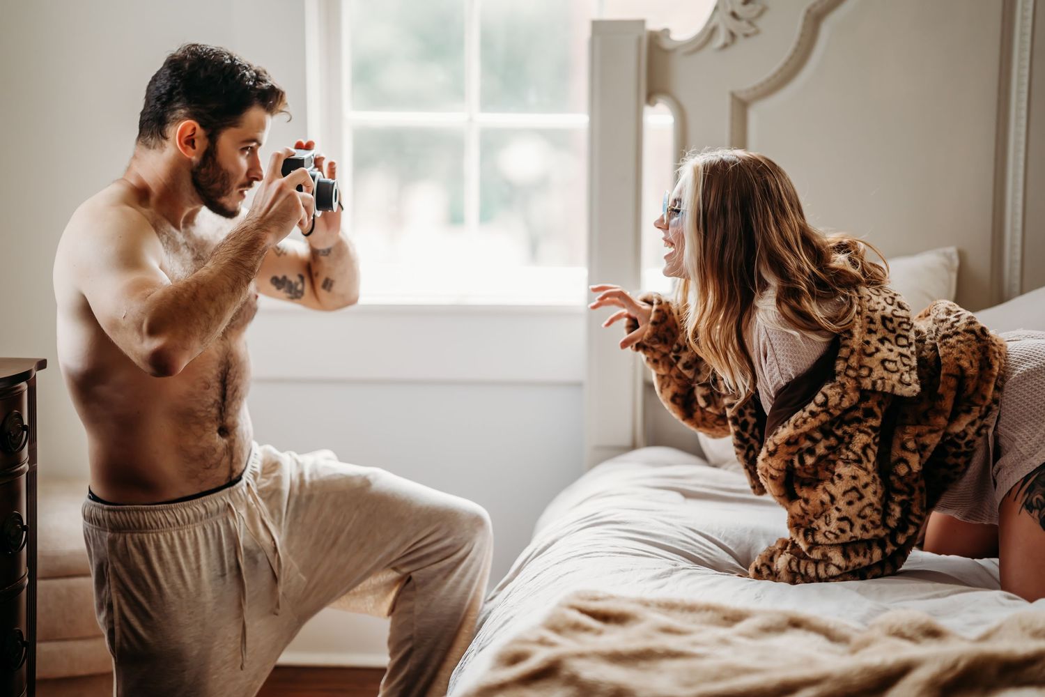 Shirtless tattooed man photographing woman in leopard coat sitting on bed.