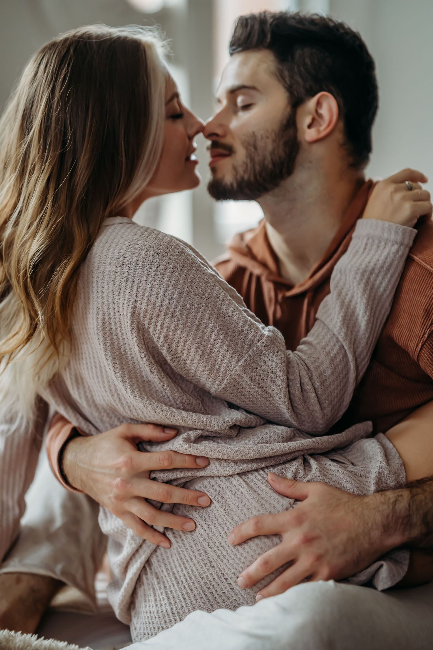 Couple sharing a tender kiss while embracing on a bed in a bright cozy bedroom.