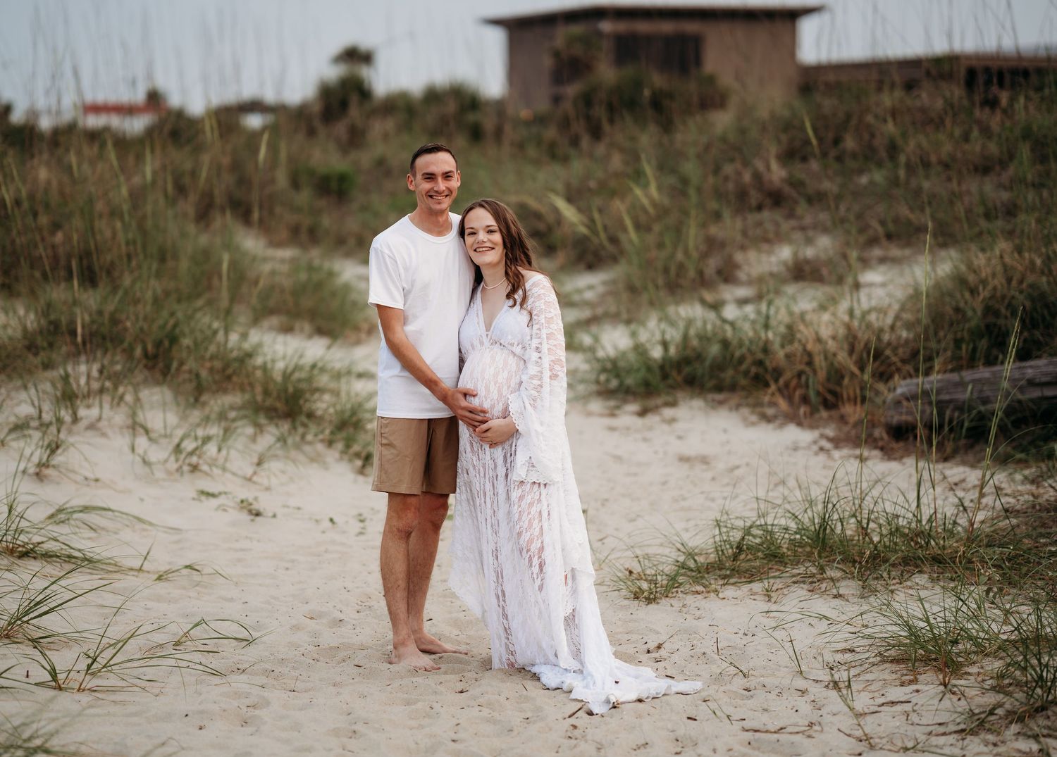 Smiling couple standing together on sandy beach dunes, woman in white lace maternity dress.