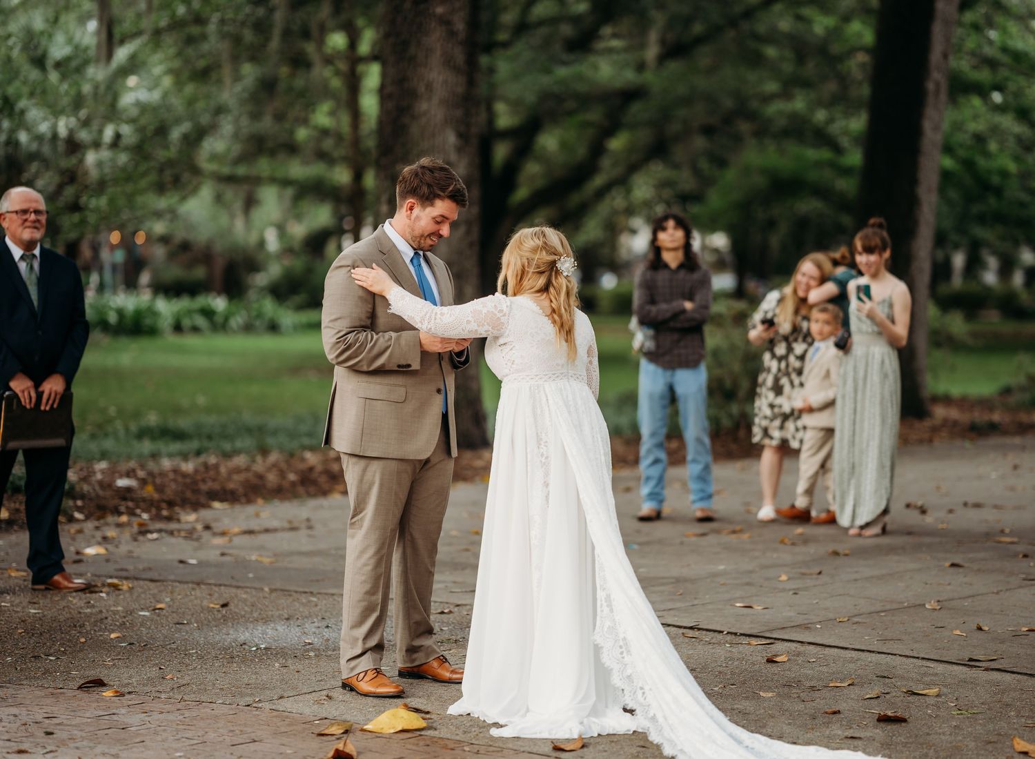 Groom in tan suit holds bride's hands during intimate outdoor wedding ceremony with guests watching.
