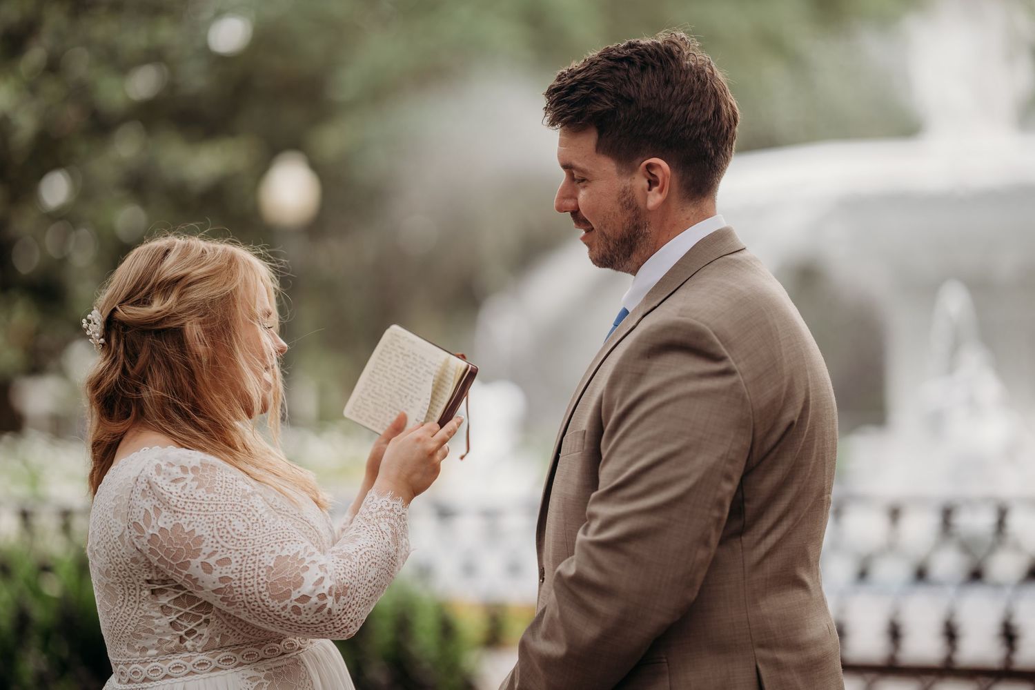 Bride in lace gown reads wedding vows from a book to her groom in a tan suit outdoors.