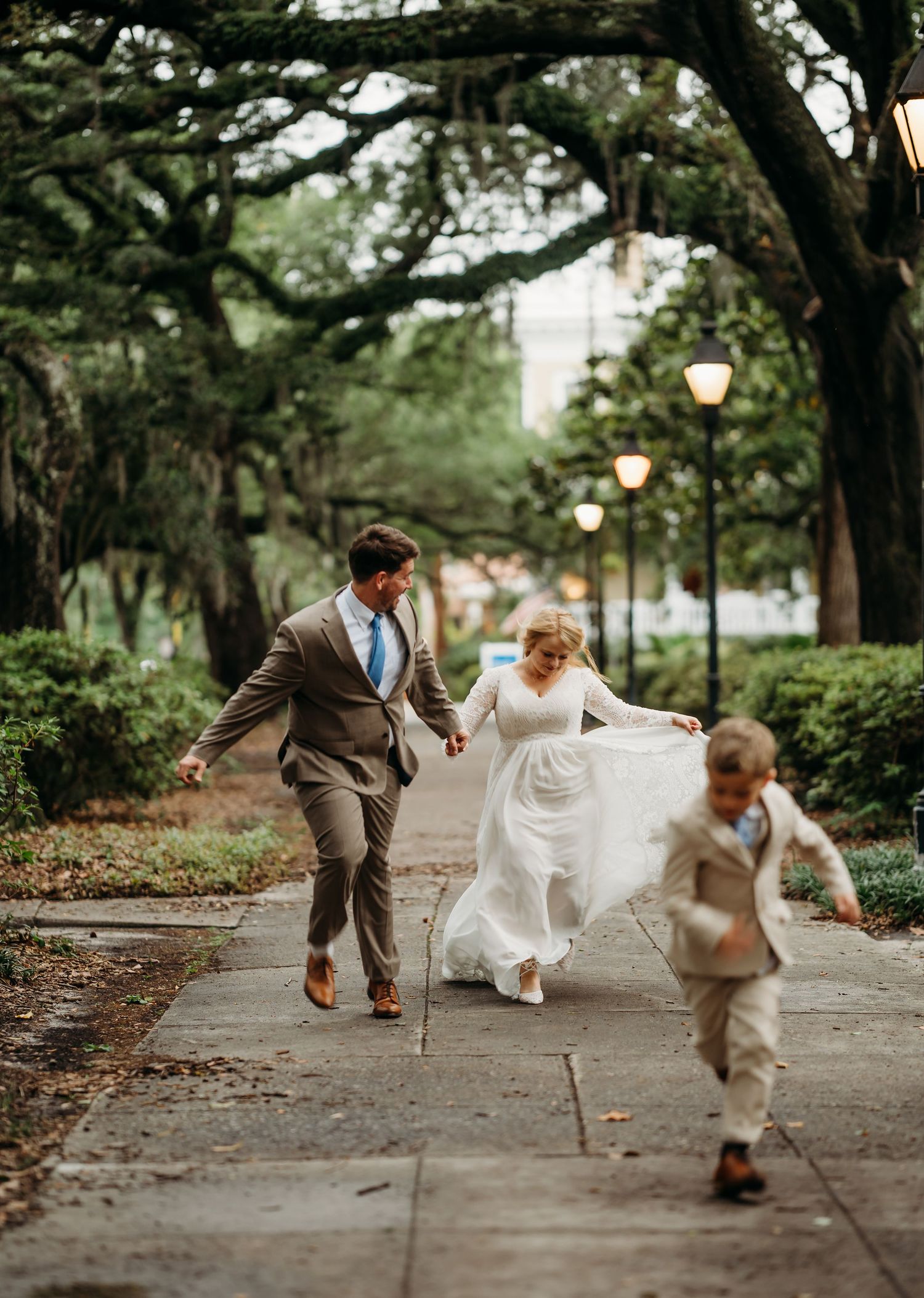 Bride and groom laugh as young boy runs ahead on tree-lined park pathway.