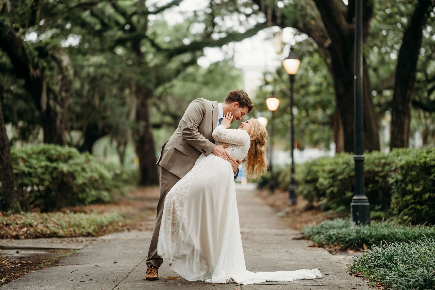 Bride and groom share a romantic dip kiss on a tree-lined sidewalk during their wedding photo session.