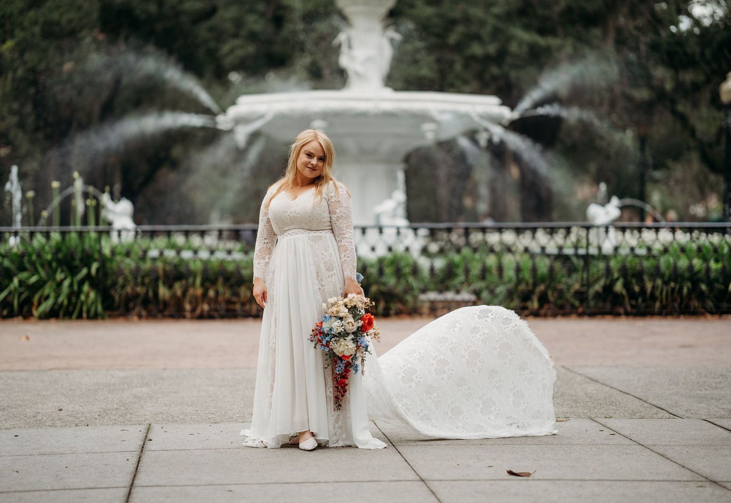 Bride in elegant white long-sleeve gown poses with colorful bouquet in front of ornate fountain.