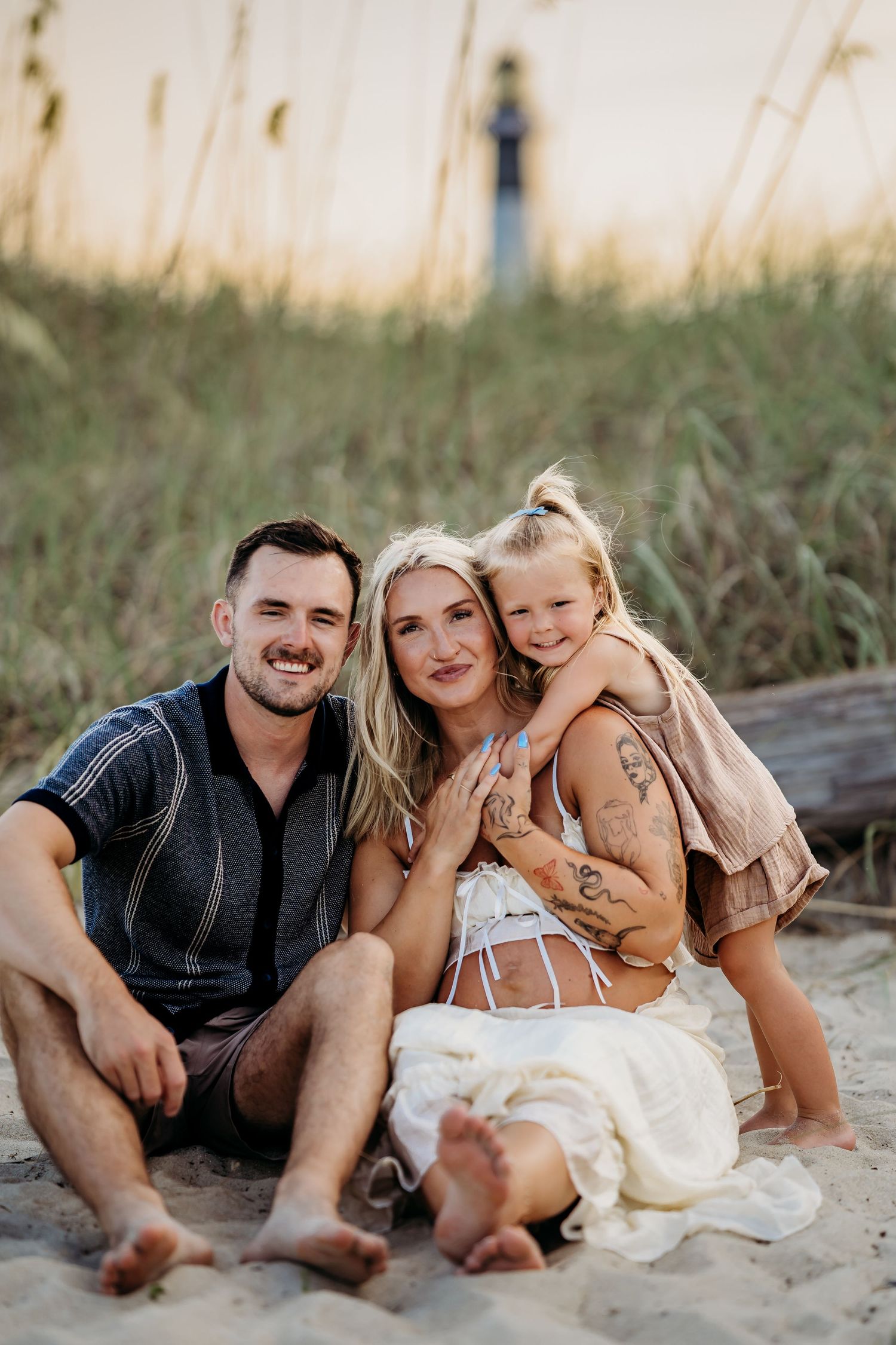 Happy family of three sits together on sandy beach at sunset near tall grass and lighthouse.