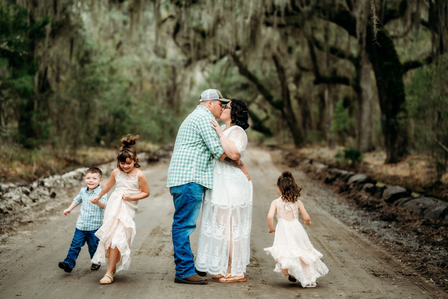 Family with two young girls in white dresses kiss on a tree-lined dirt road.