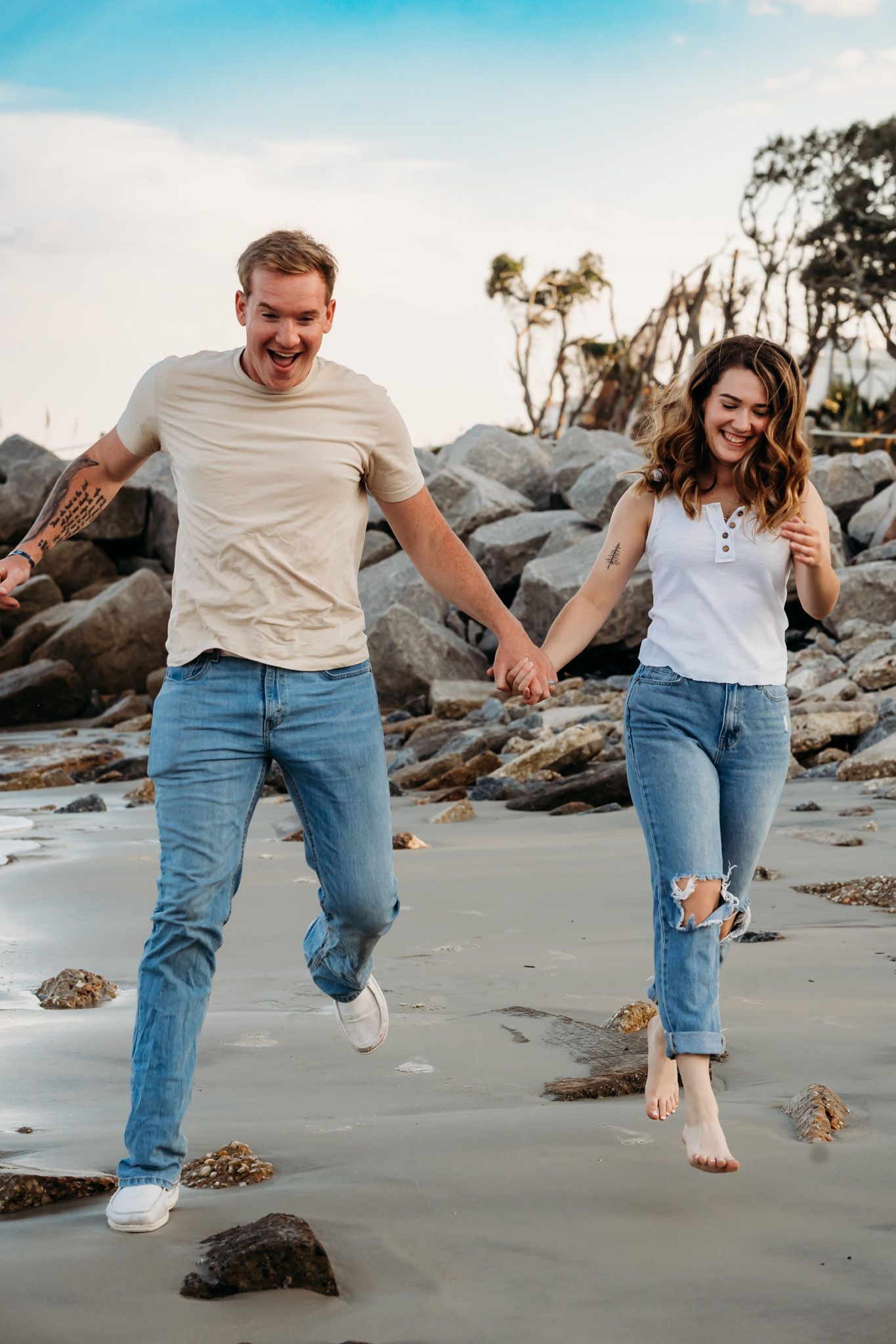 A joyful couple holds hands and runs along a rocky beach, both wearing jeans and casual tops under a blue sky.