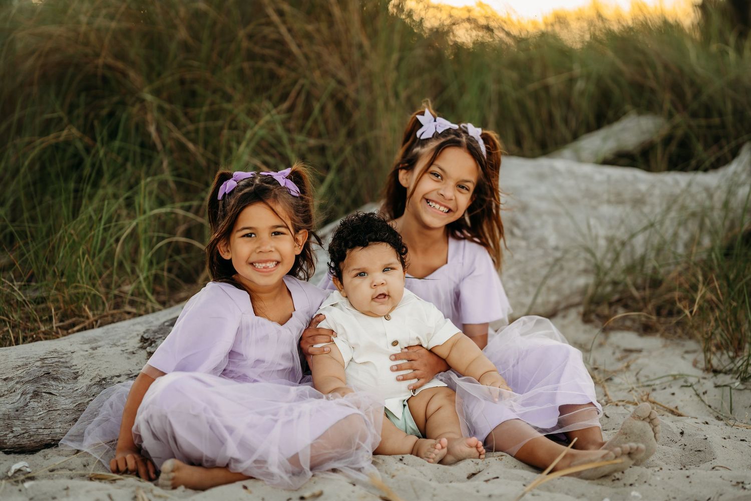 Two smiling girls in lavender dresses sit on sandy beach with baby sibling, driftwood and tall grass behind them.