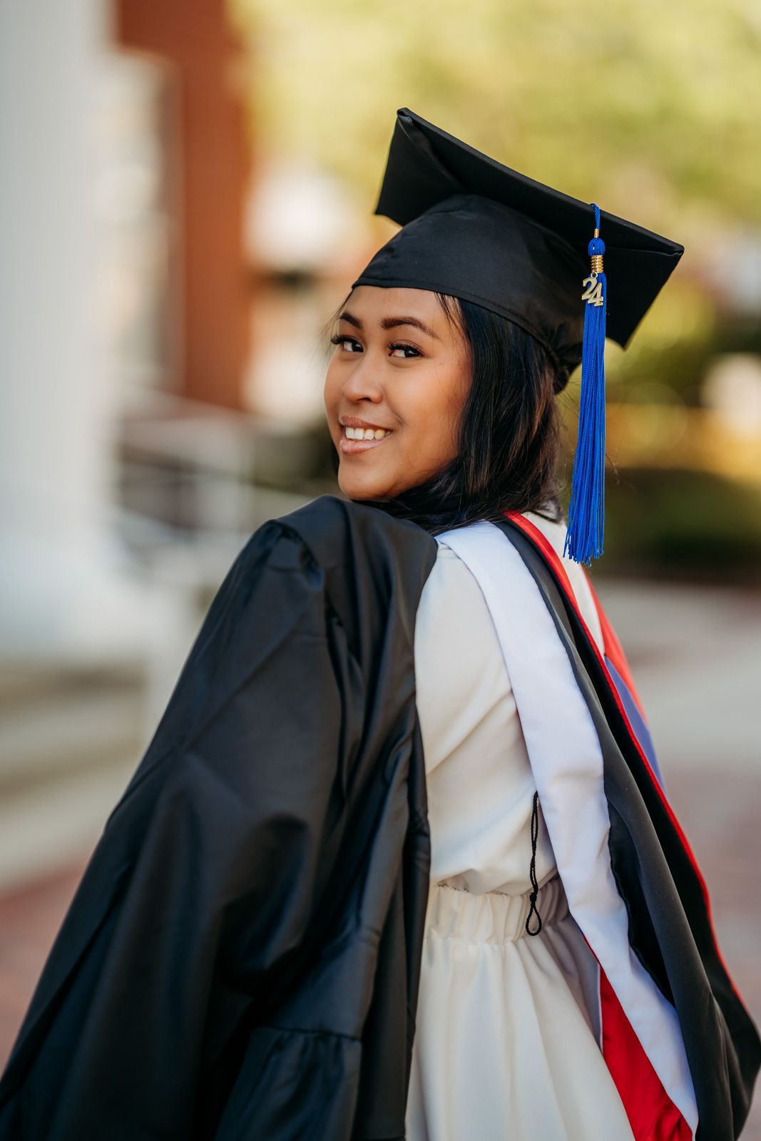 Smiling young woman in black graduation cap and gown with blue tassel and red sash outdoors.