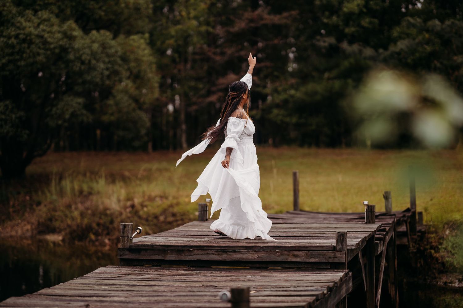 Woman in white dress jumping joyfully on wooden dock surrounded by lush green trees and pond.