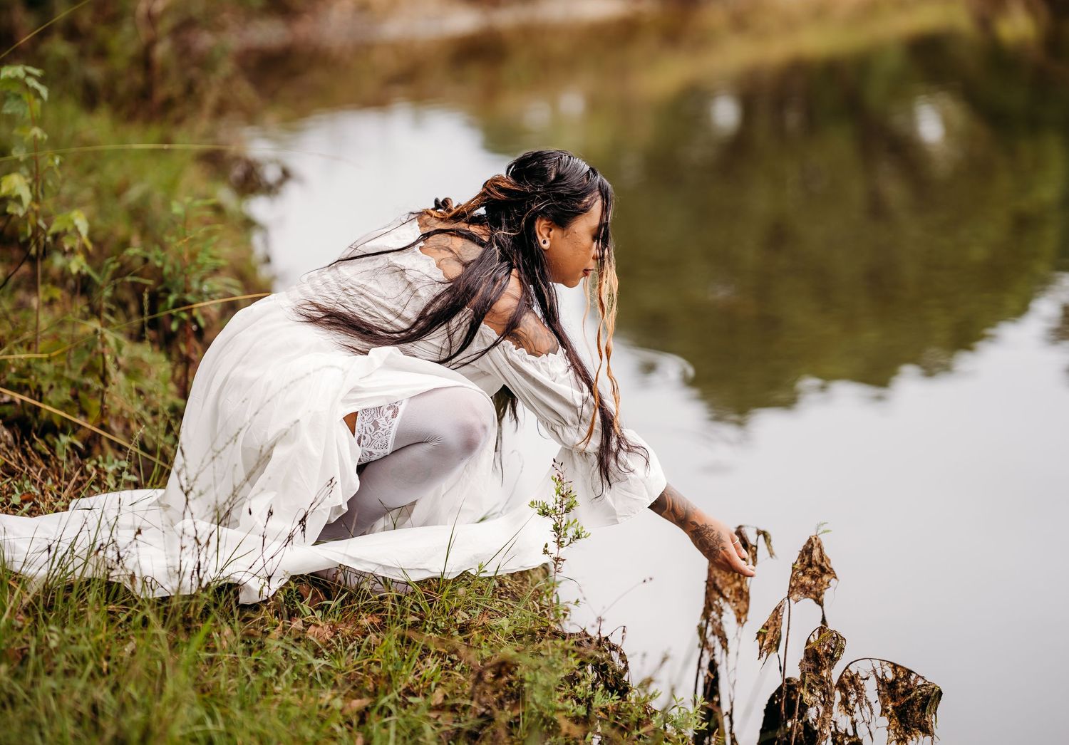 Woman in white dress and tights crouching by a calm pond reaching toward the water.
