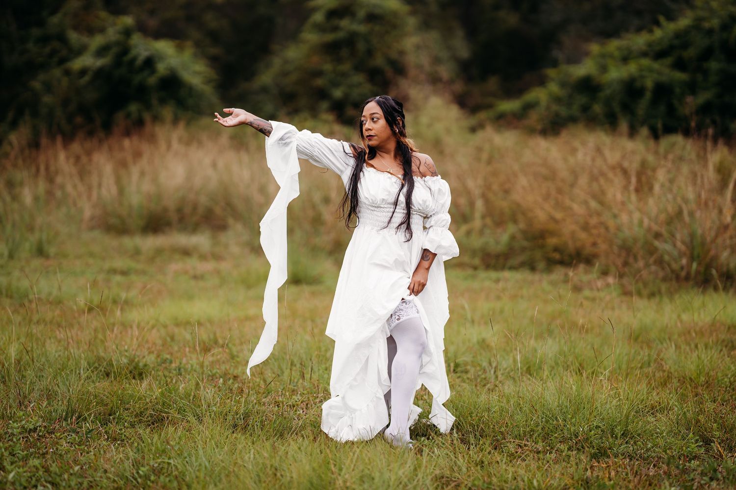 Woman in white off-shoulder dress posing expressively in grassy field with fabric flowing.