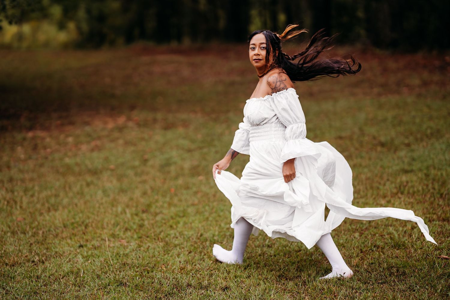 Young woman in flowing white dress running dynamically across green field with trees behind.