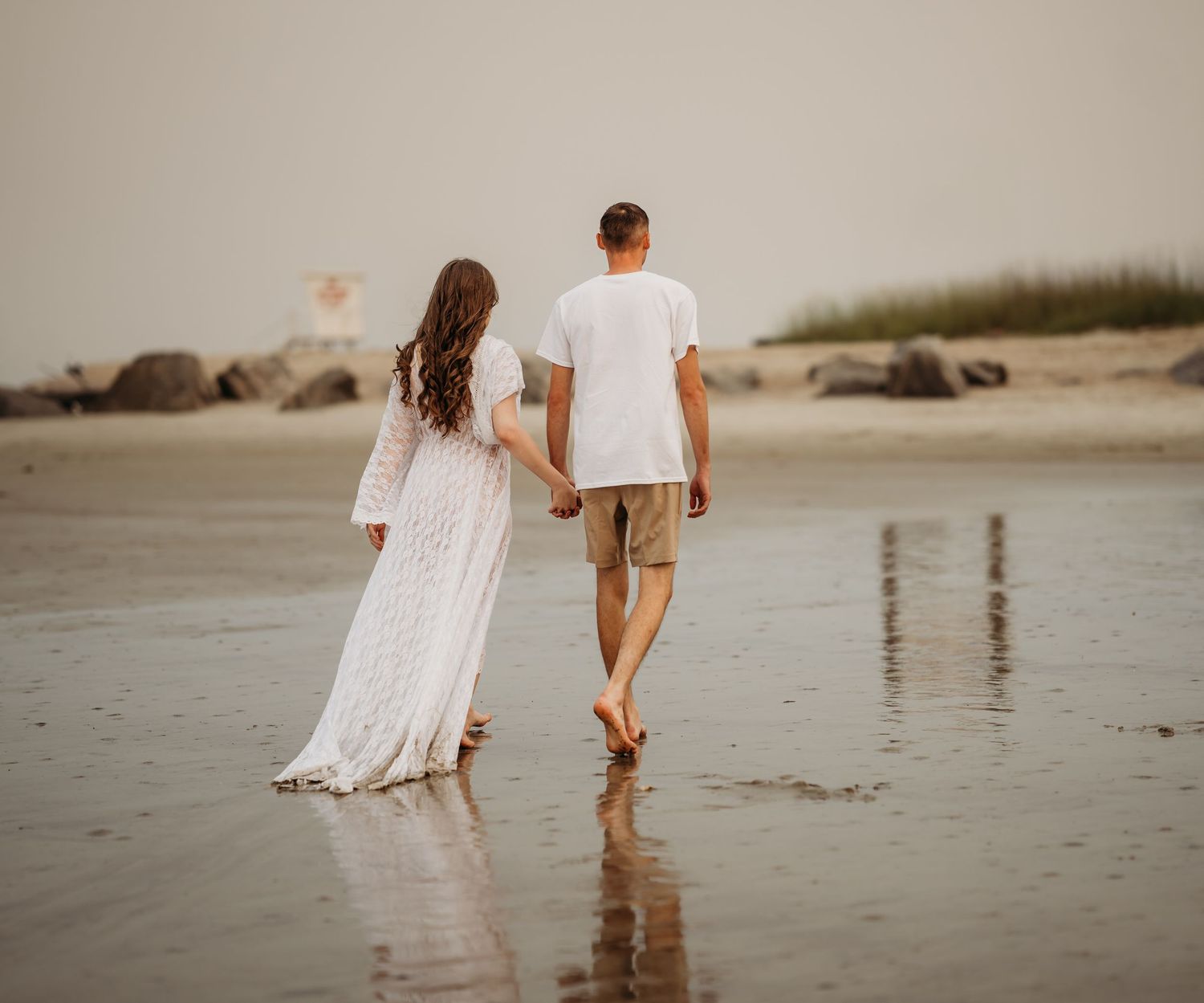 Pregnant couple walks hand in hand along wet shoreline during beach maternity photoshoot at dusk.