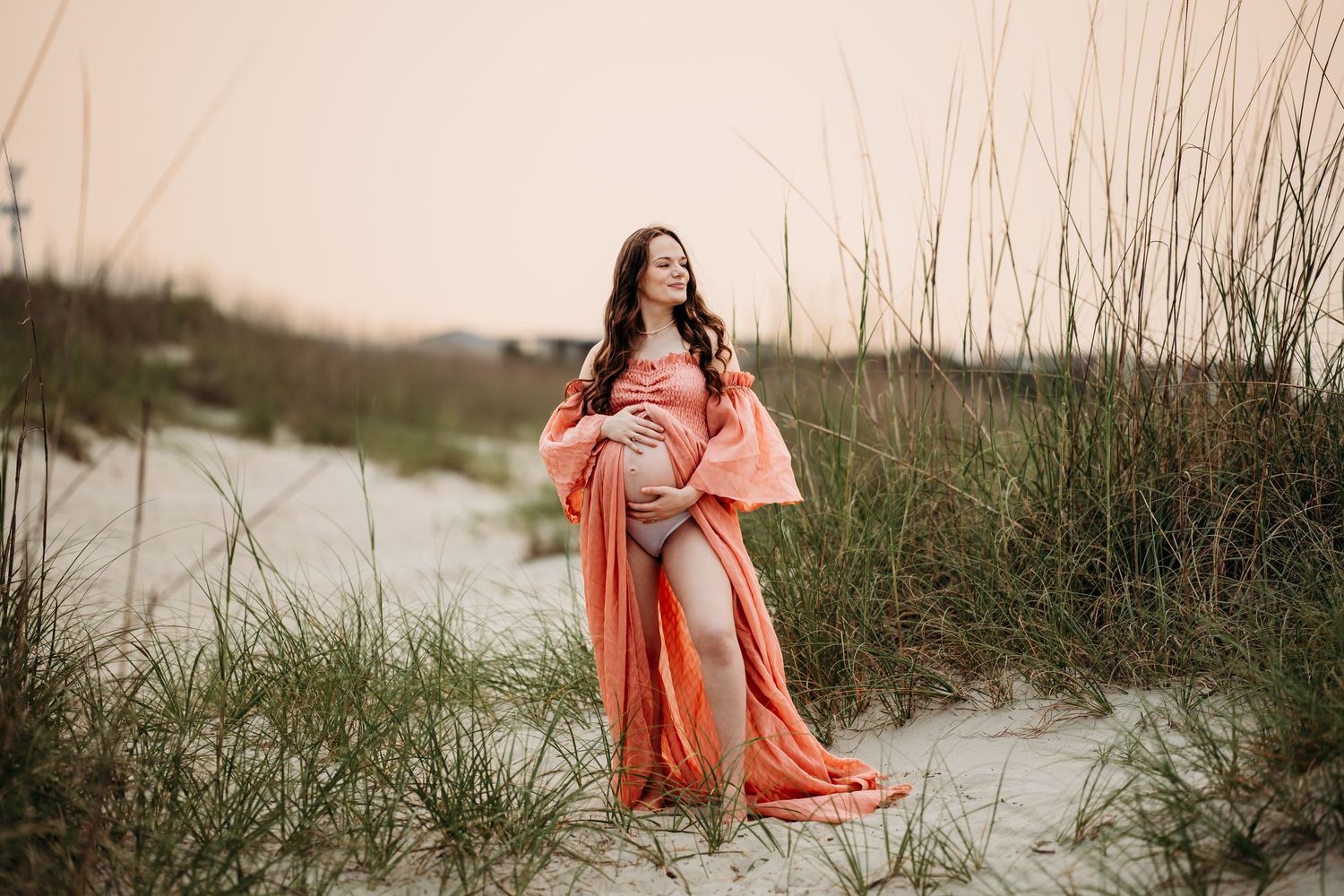 Pregnant woman in elegant coral flowing dress cradling baby bump on sandy beach at golden hour.